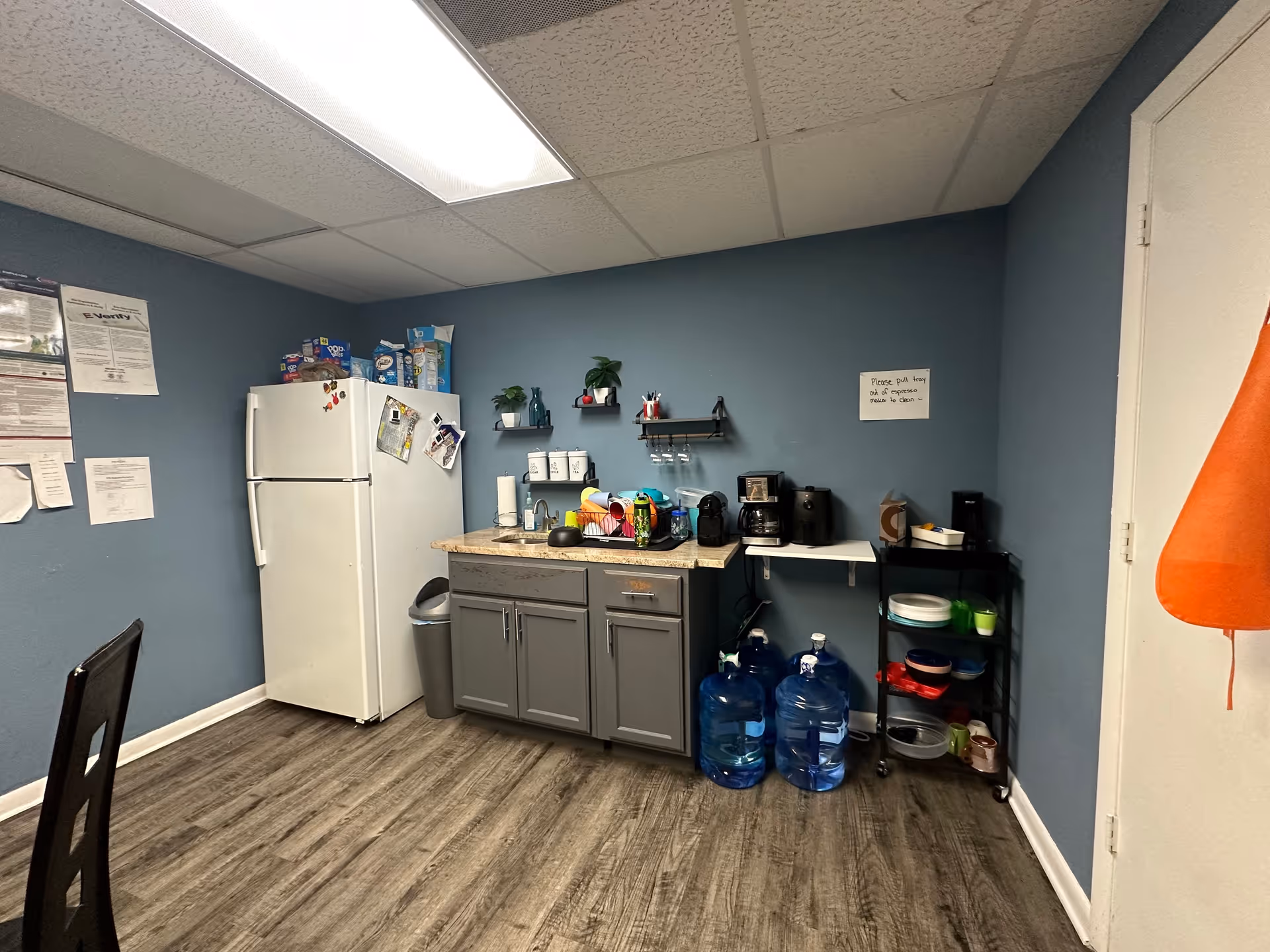 Small blue-walled kitchenette with fridge, cabinets, counter, coffee station, and water jugs.