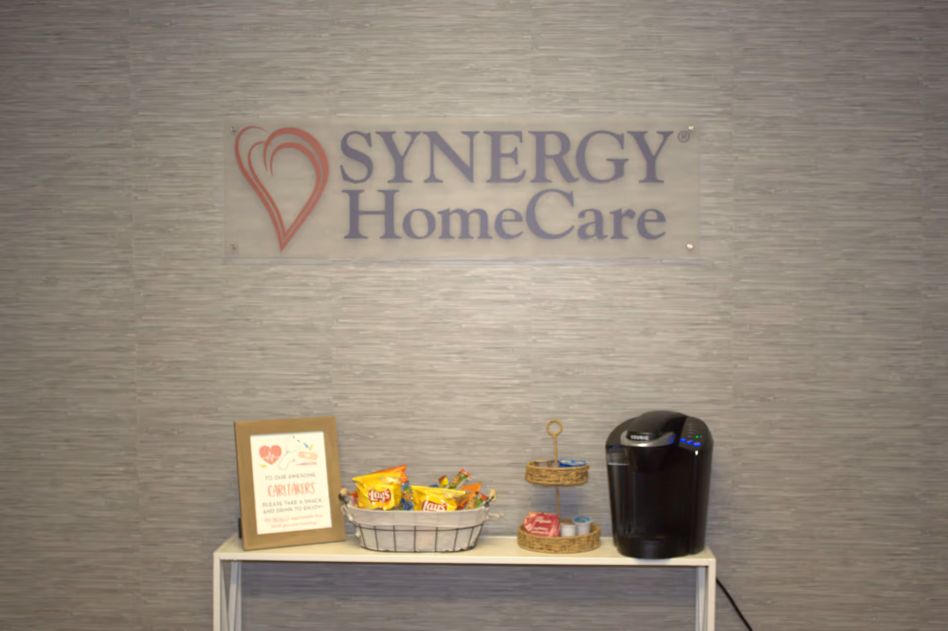Interior wall with SYNERGY HomeCare sign, snack basket, and coffee station on a table.