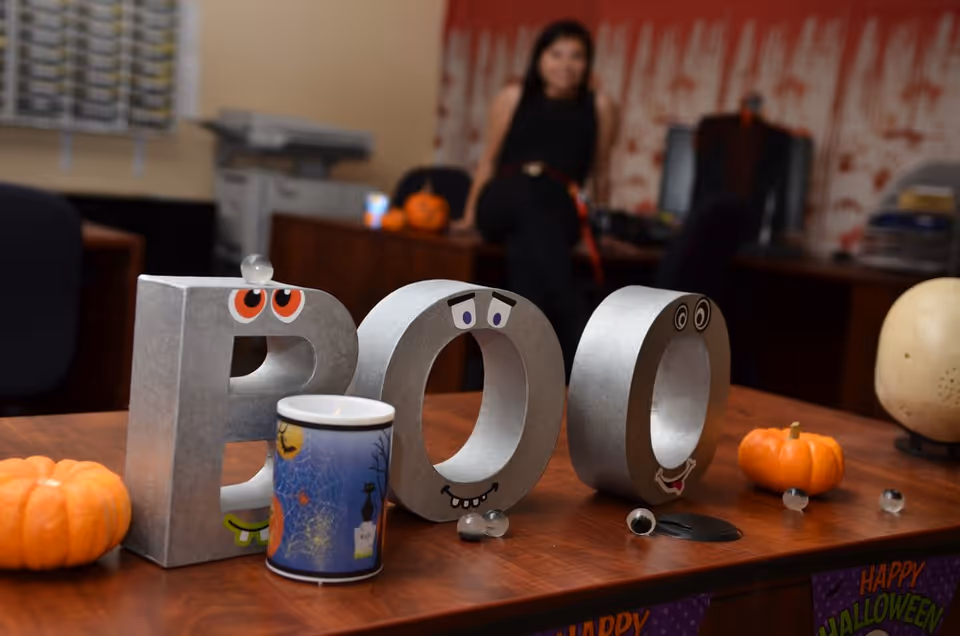 Metal letters 'POO' with googly eyes on a table; pumpkins and mug; blurred person in background.