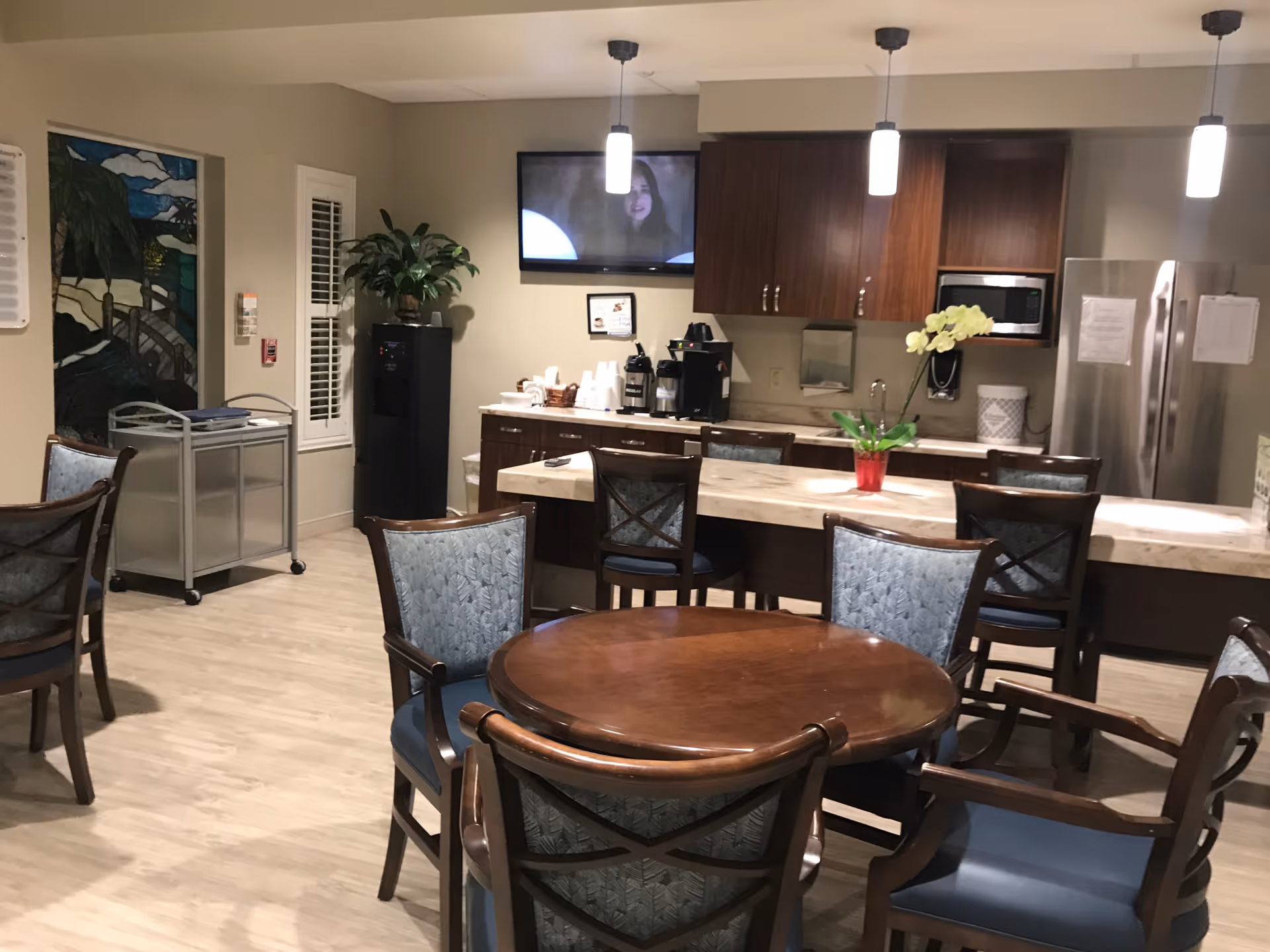 Indoor senior living lounge with a round wooden table, blue chairs, and coffee station.