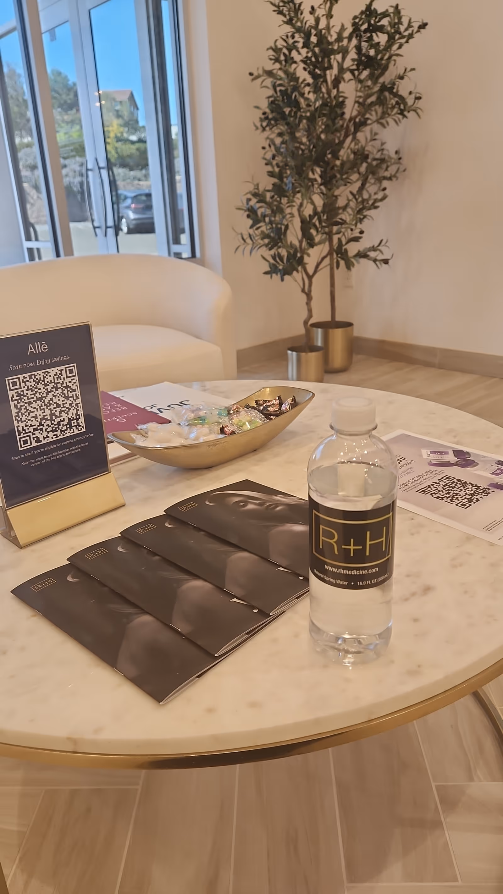Marble round table with brochures, a water bottle, and a tall plant in a bright lobby.