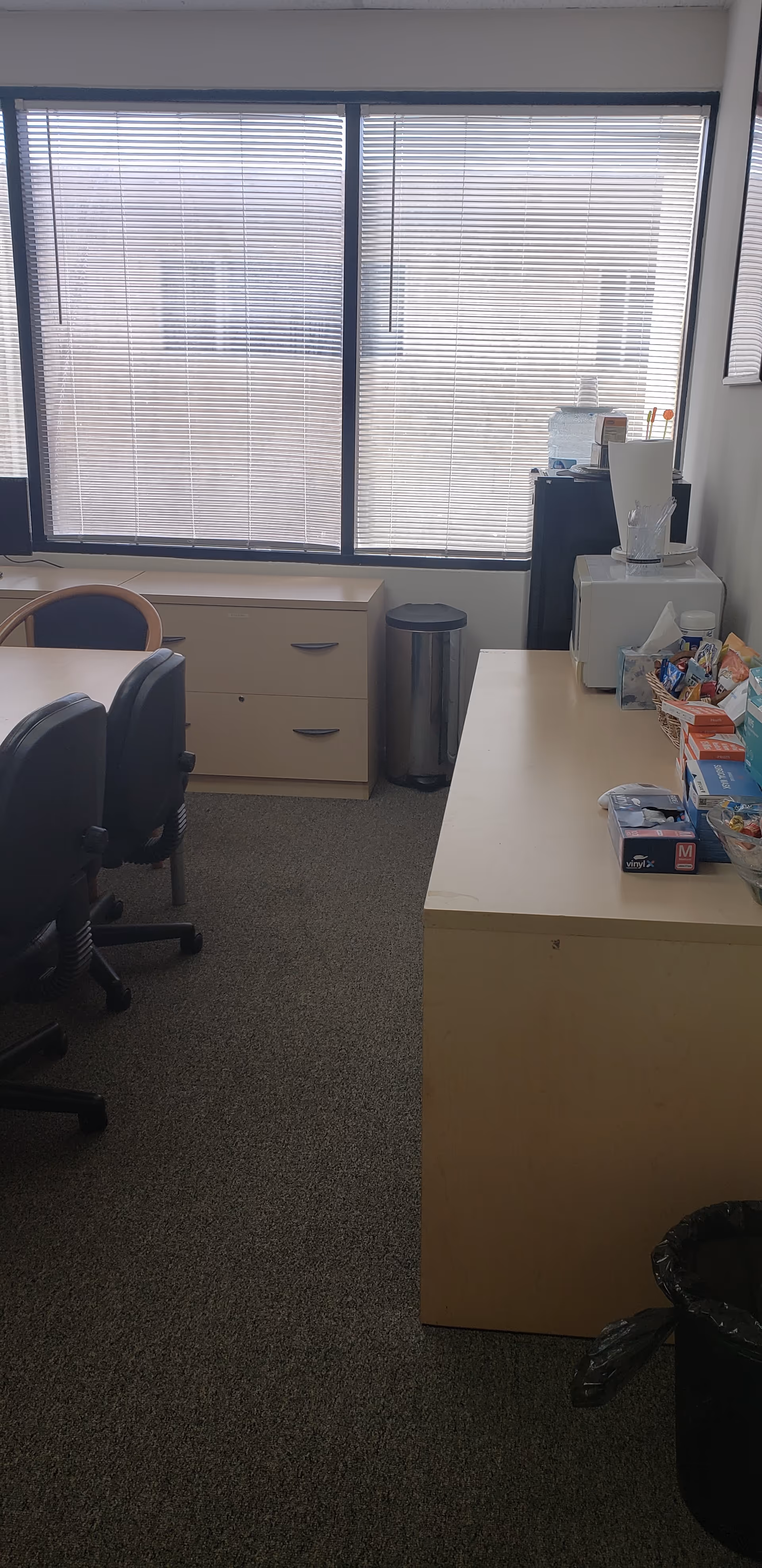 Office kitchenette area with chairs, a desk, large window blinds, and snacks on the counter.