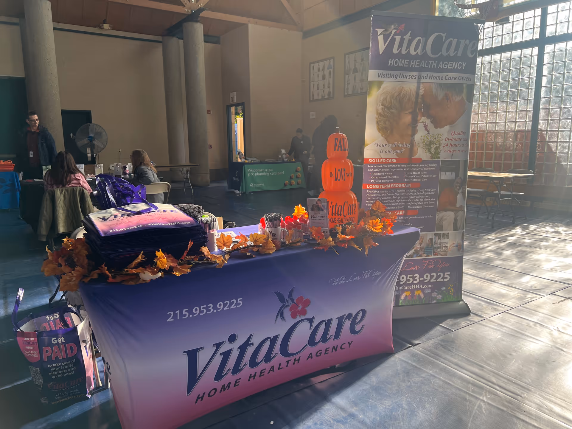 Indoor hall with VitaCare promotional booth, banner, autumn decorations, and visitors.