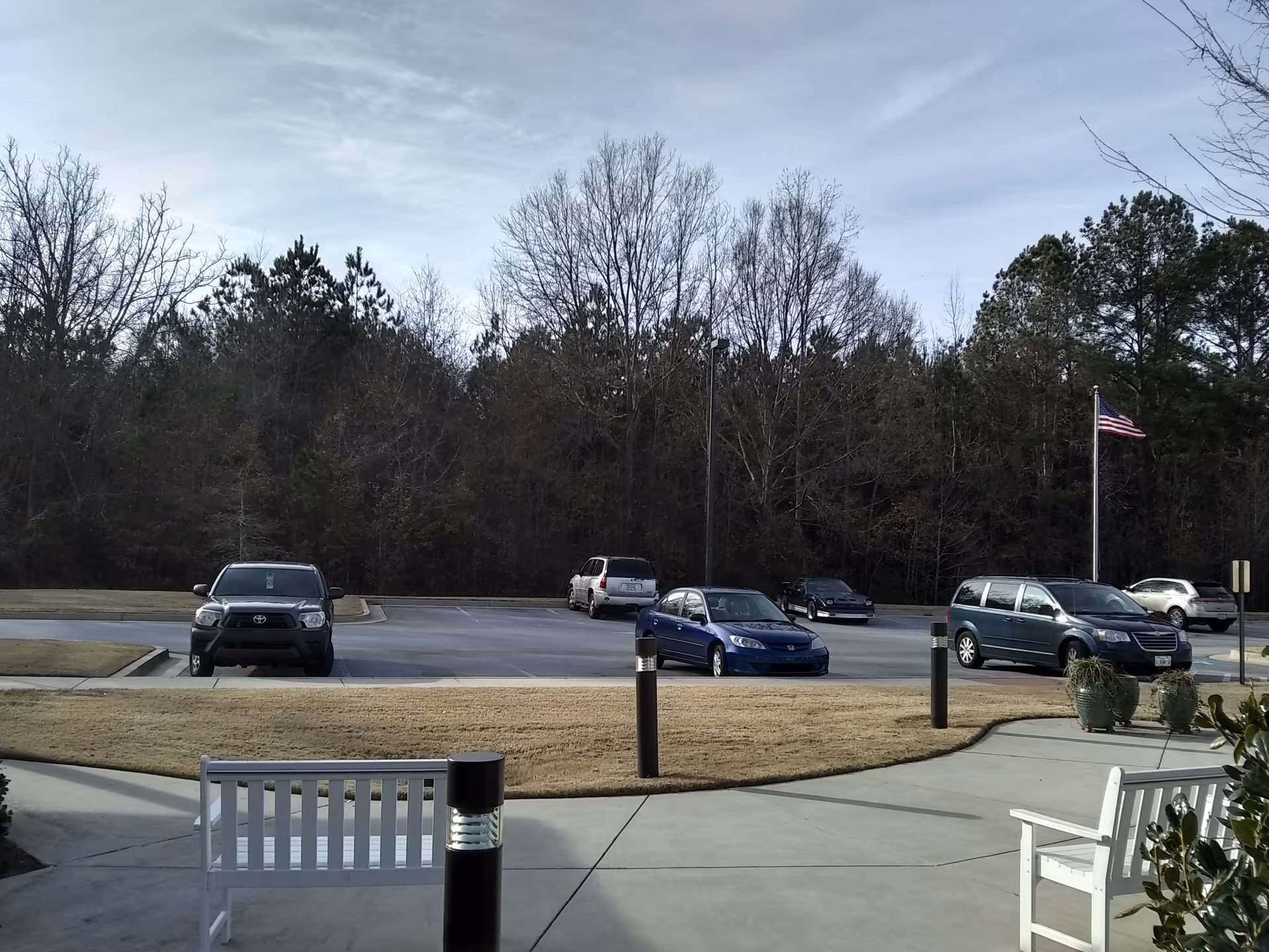 A parking lot with several cars, trees in background, and white outdoor benches near entrance.
