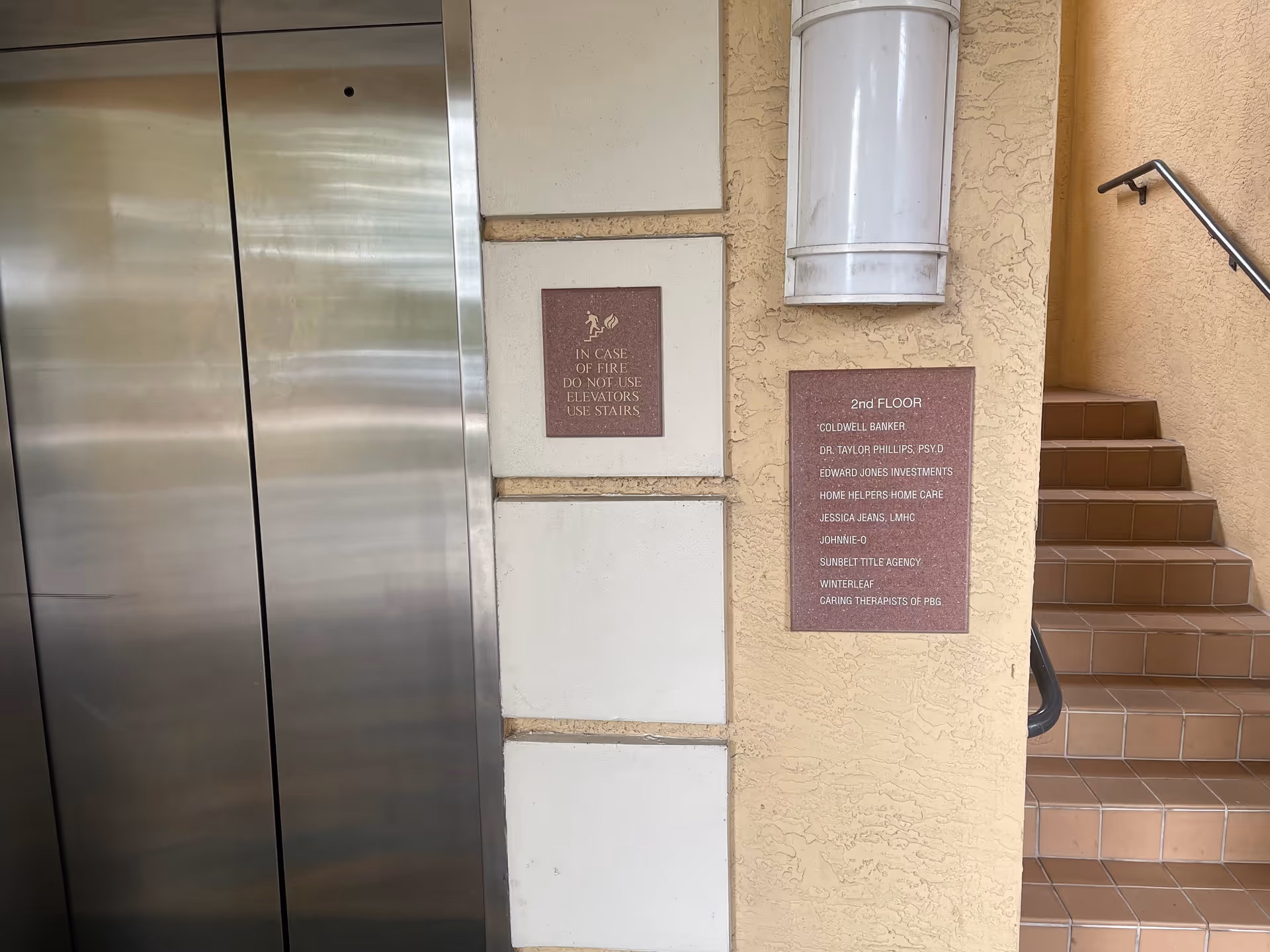 Elevator doors on left, beige walls with plaques, and brown tiled stairs on the right.
