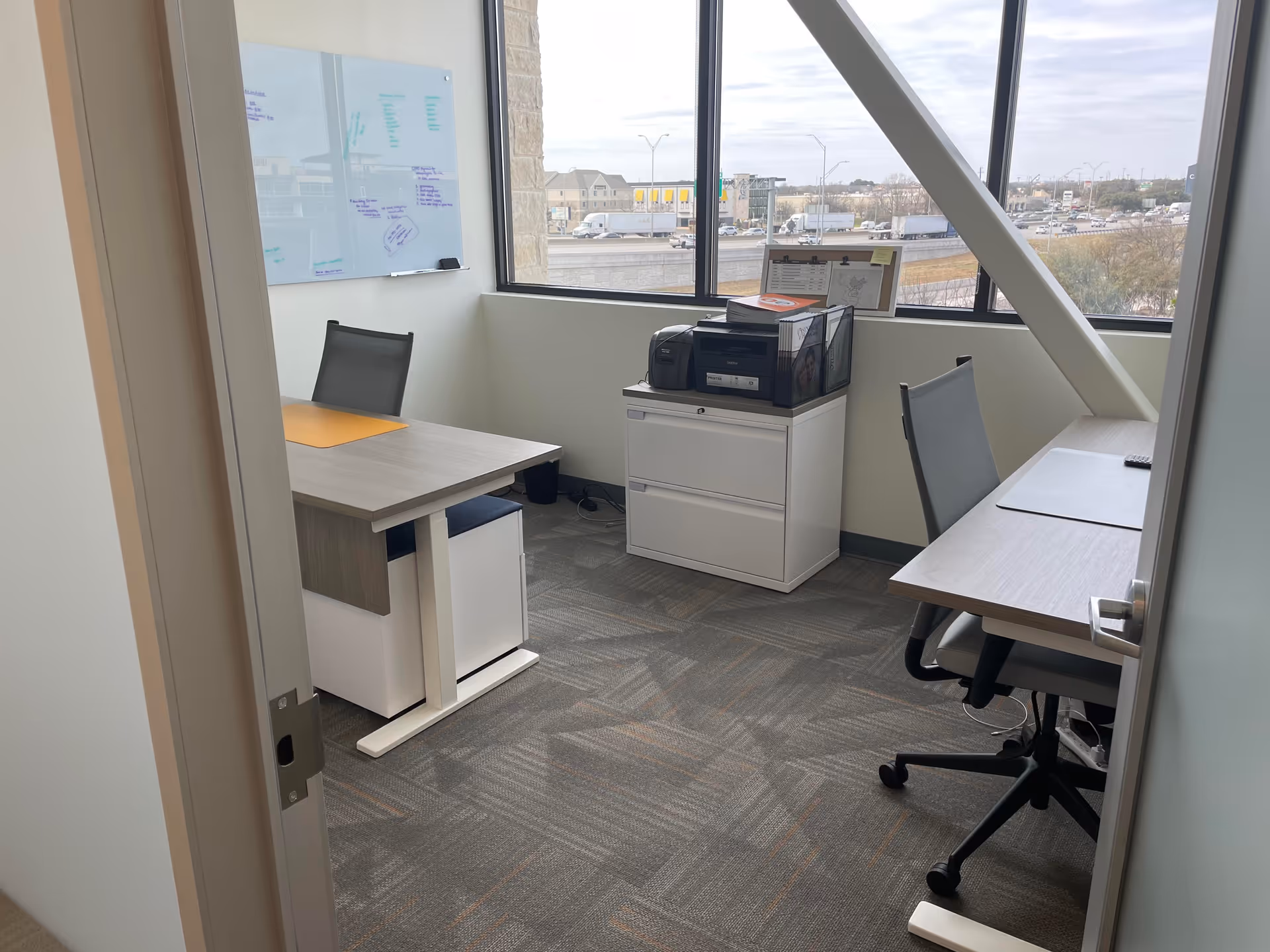 Small office space with two desks, chairs, printer, and a whiteboard by large windows.