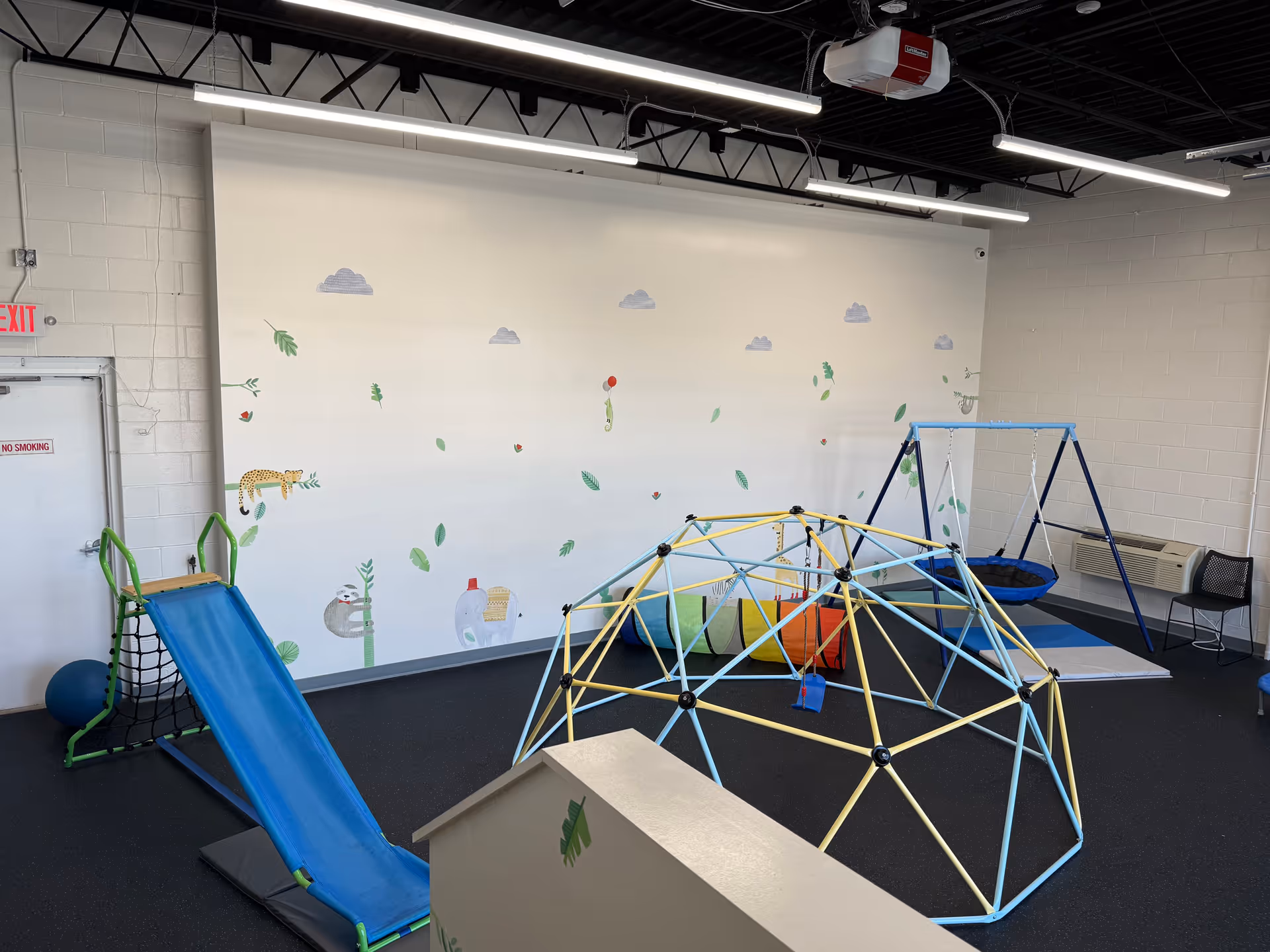 Indoor therapy playroom with climbing dome, blue slide, and leafy wall mural.