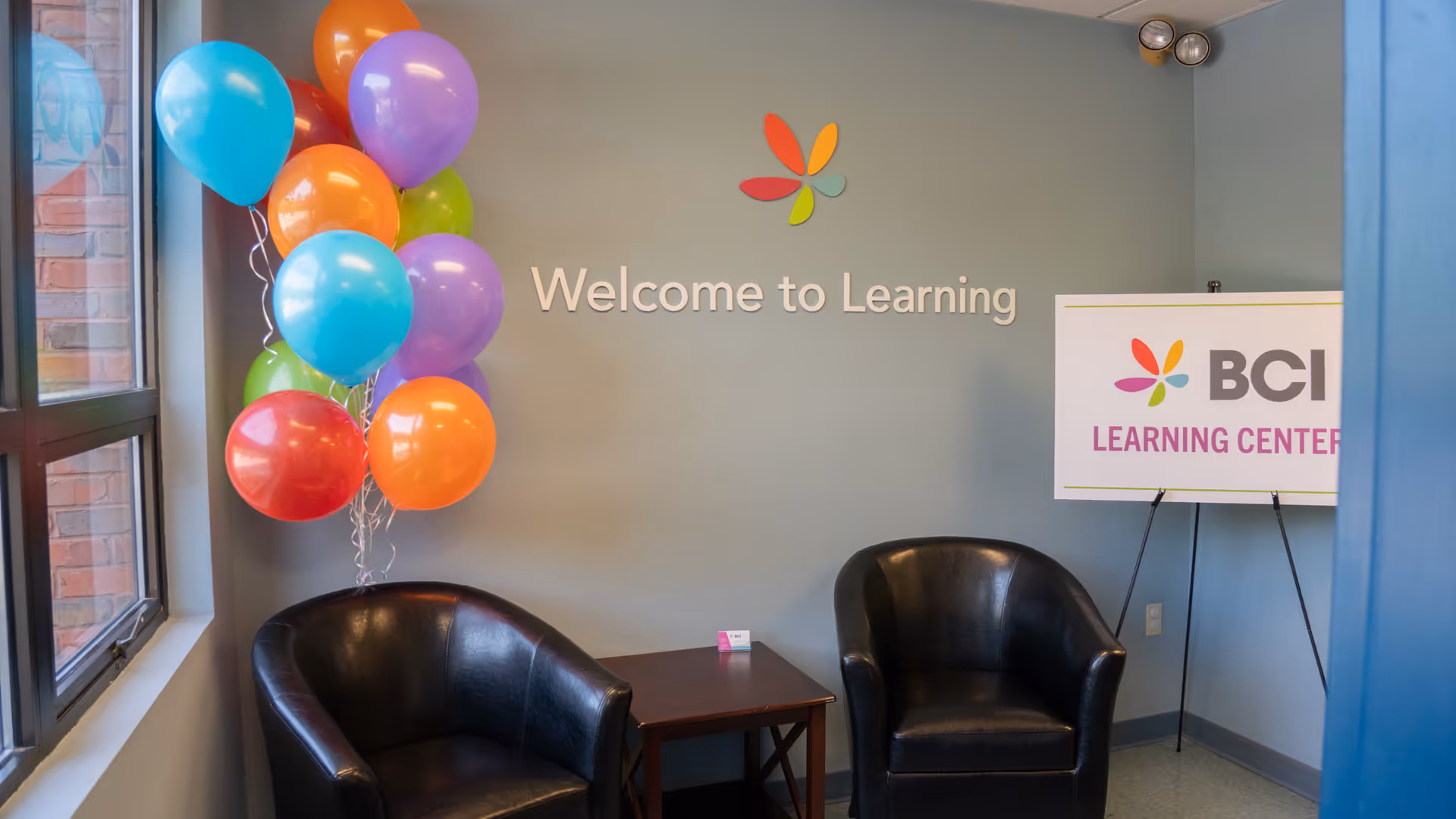 Lobby area with two black chairs, a small table, balloons, and 'Welcome to Learning' sign.