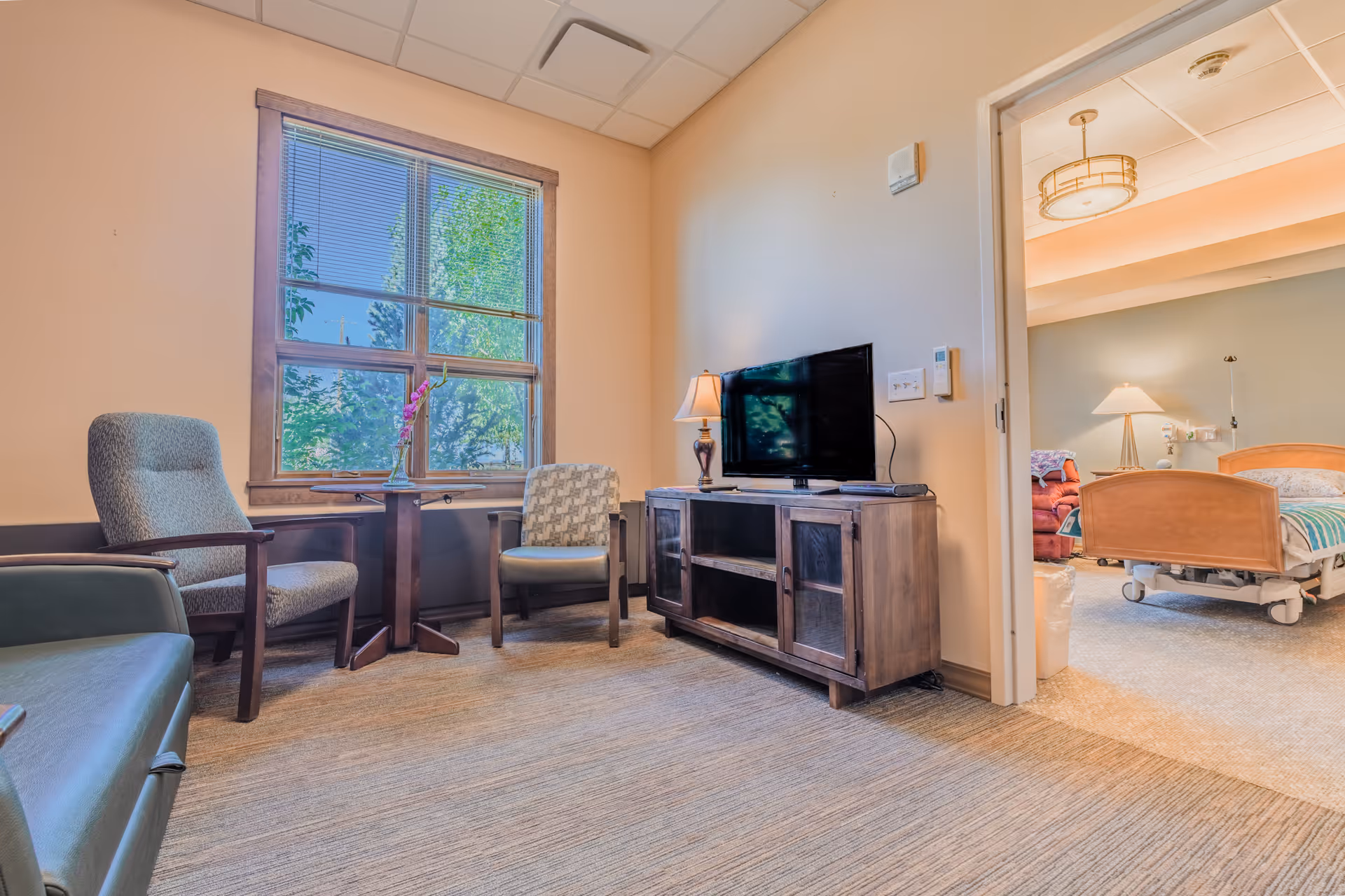 Cozy living room lounge with chairs, window view, TV, and adjoining patient room.