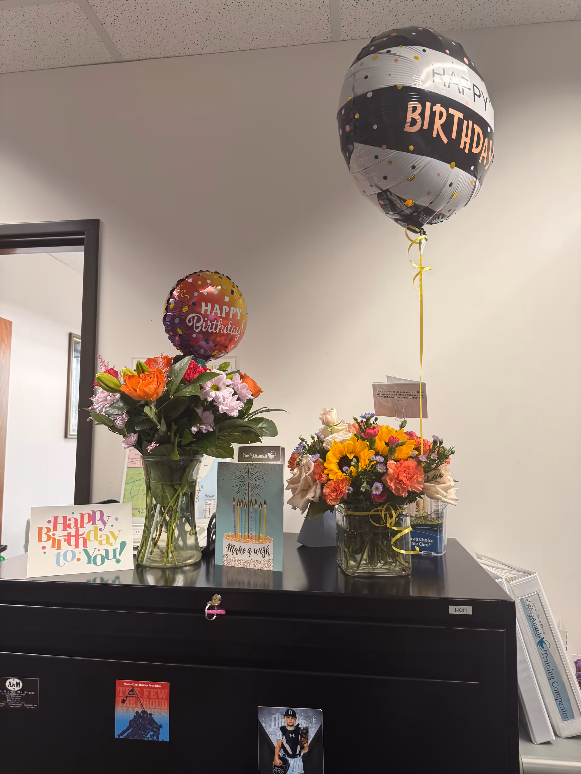 Birthday balloons and flowers decorate a dark cabinet in a senior care facility.