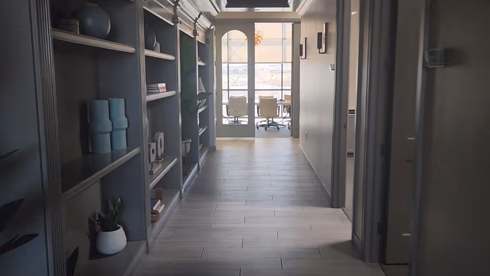 Dim hallway with built-in shelves on the left and a distant seating area beyond glass doors.