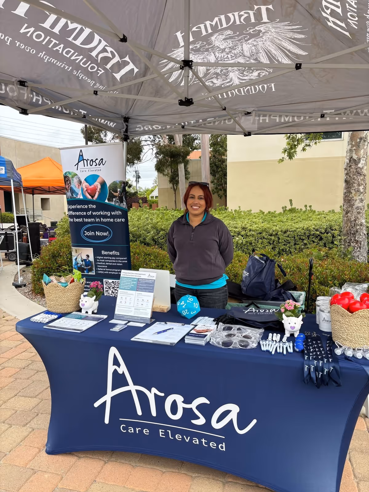 A staff member stands at an Arosa booth under a large white canopy outdoors.