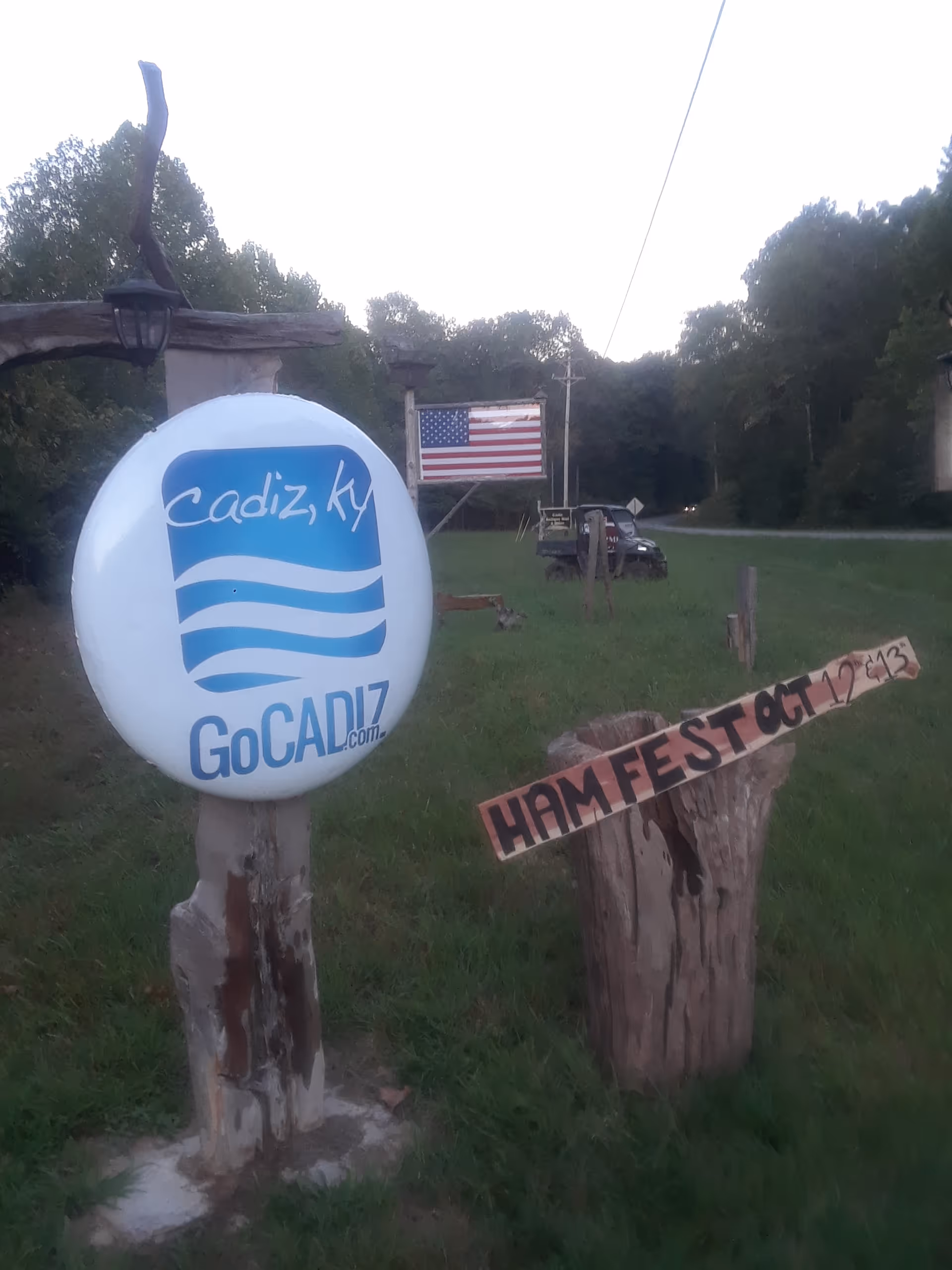 Outdoor rural scene with Cadiz GoCadiz sign, American flag, and ham festival sign.