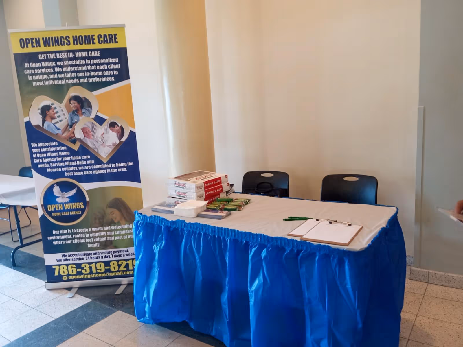 Promotional table with blue skirt and Open Wings Home Care banner in an interior lobby.