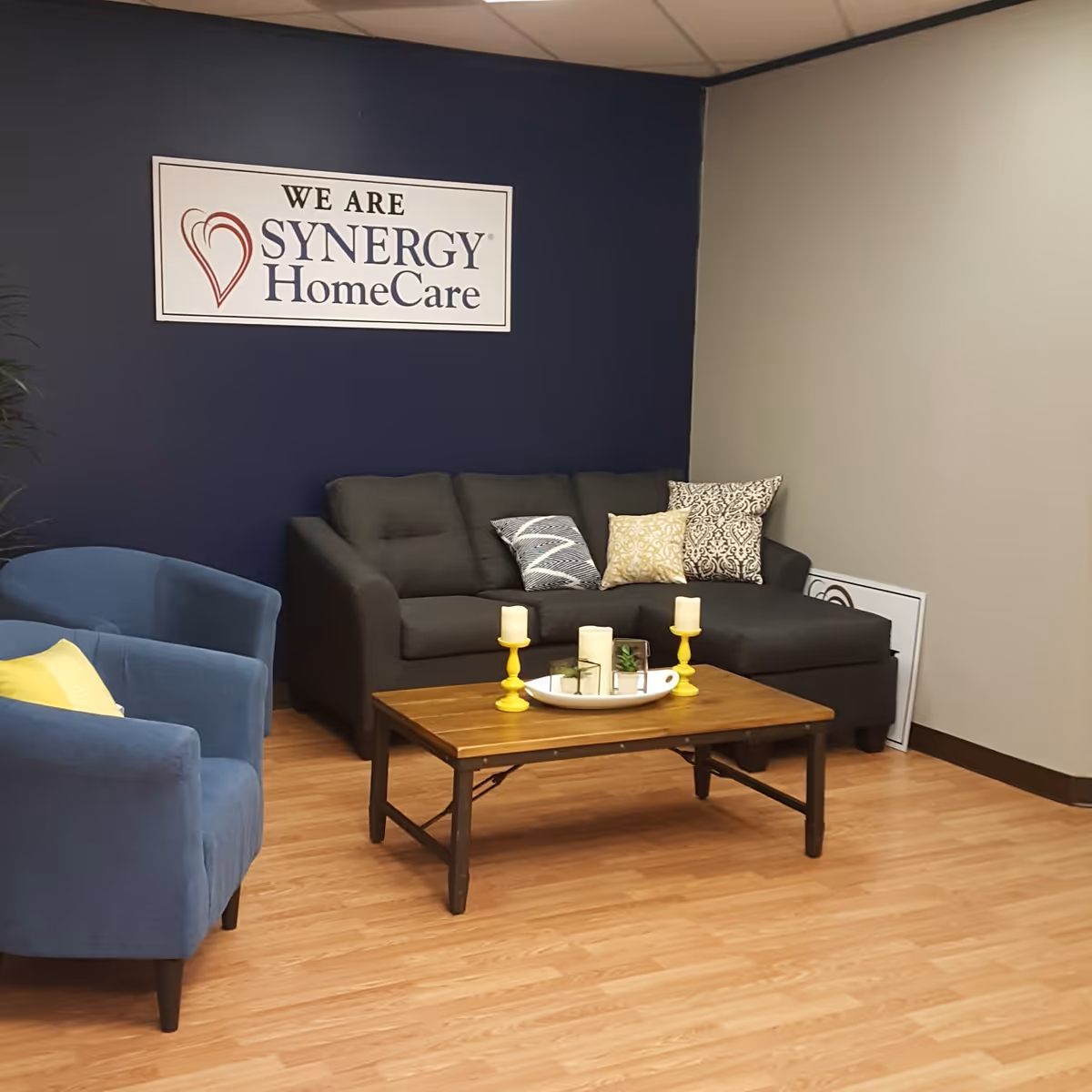Lounge area with blue chairs, a gray sofa, coffee table, and a SYNERGY HomeCare sign.