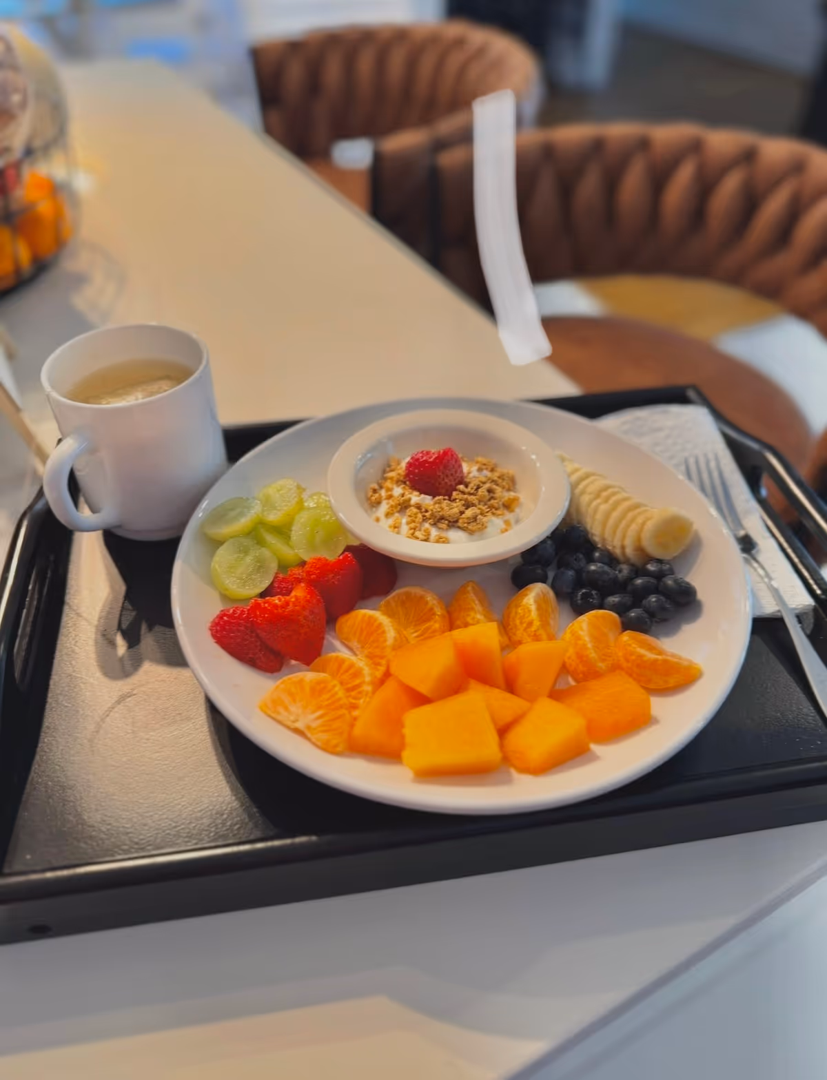 Fruit platter with grapes, berries, orange slices, mango, yogurt, granola, banana, and coffee on a tray.