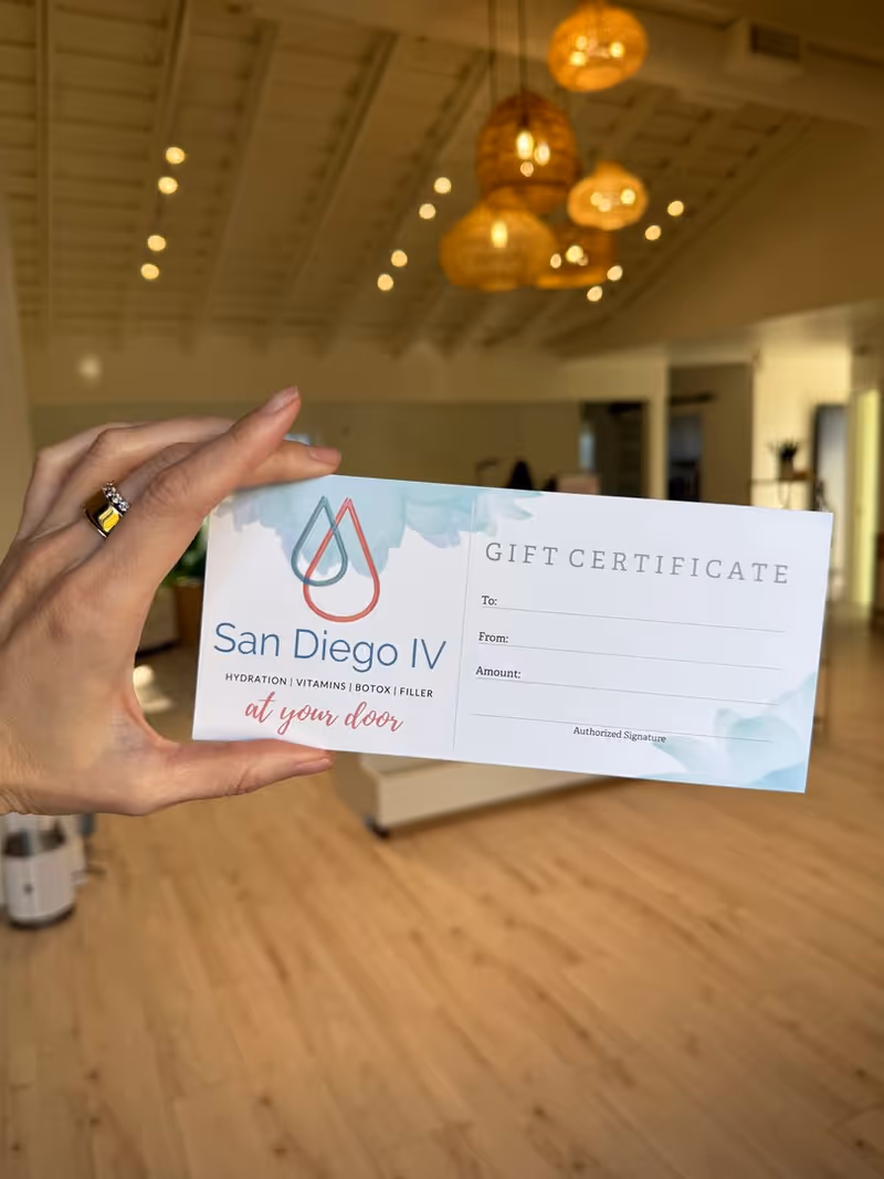 Hand holds San Diego IV gift certificate in a bright, airy lobby with wooden floor.