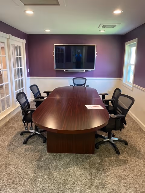 A conference room with a large wooden table, chairs, and a mounted TV on purple wall.