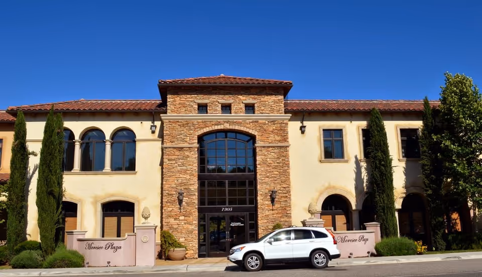 Stone and beige senior living facility facade with arched windows and a parked white SUV.