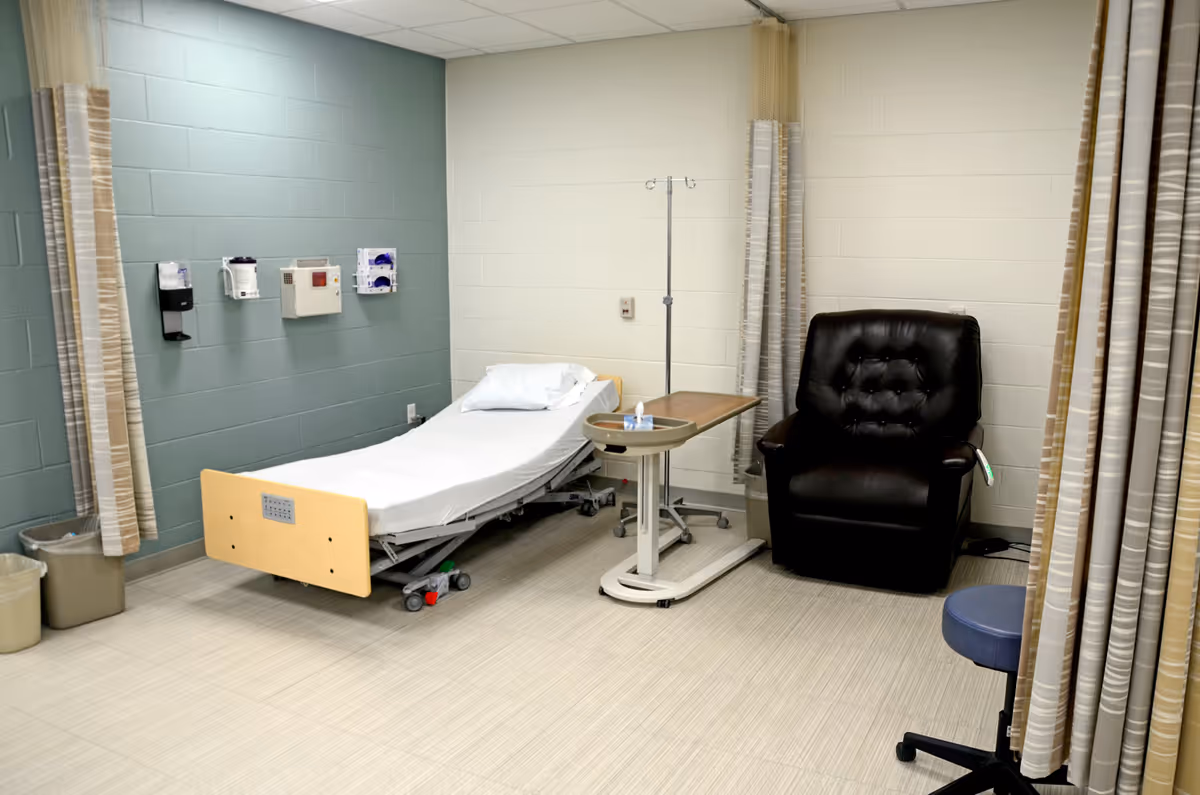 Hospital-style patient room with a bed, IV pole, table, chair, and curtains.