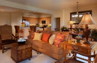 Cozy living room with sofa, chairs, lamps, and an open kitchen in the background.