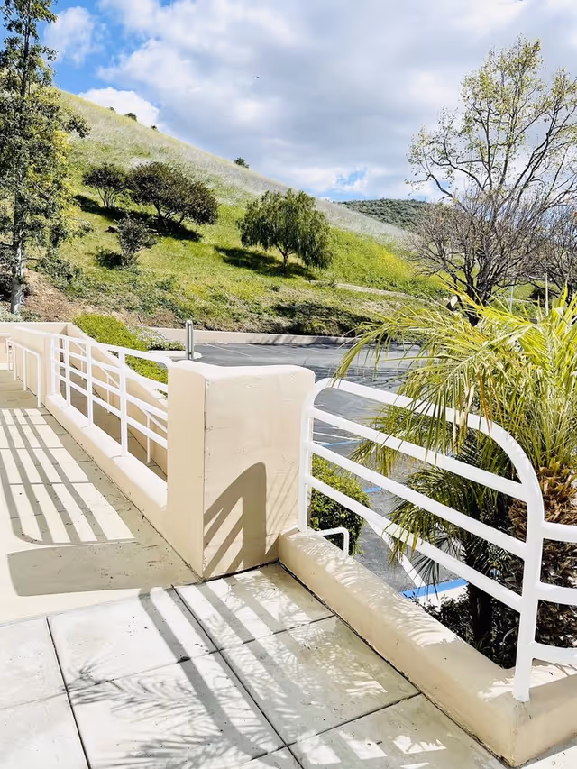 Sunny outdoor walkway with white railing, concrete patio, and grassy hillside in the background.