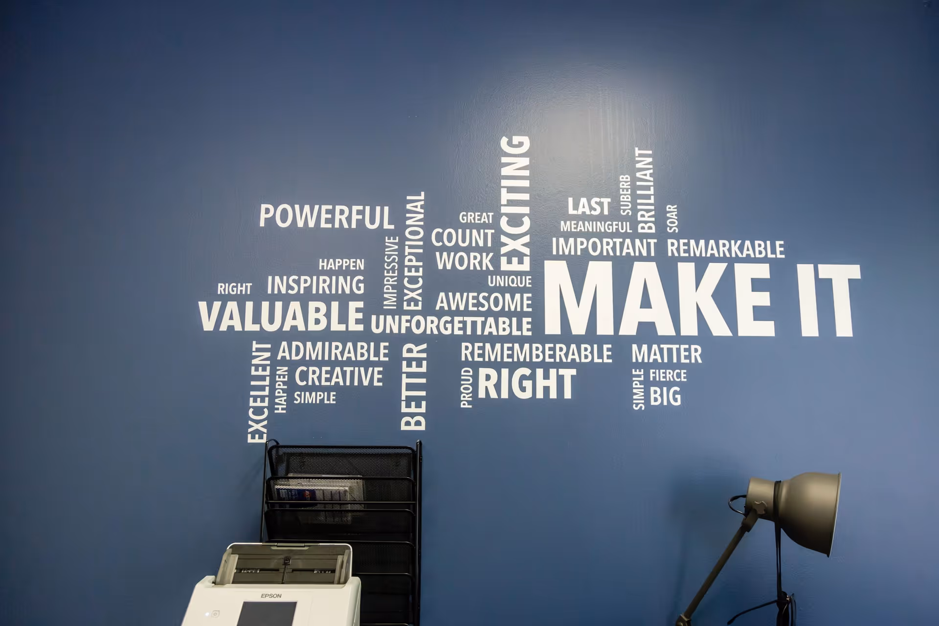 Blue wall features large white motivational words with a printer and office lamp.