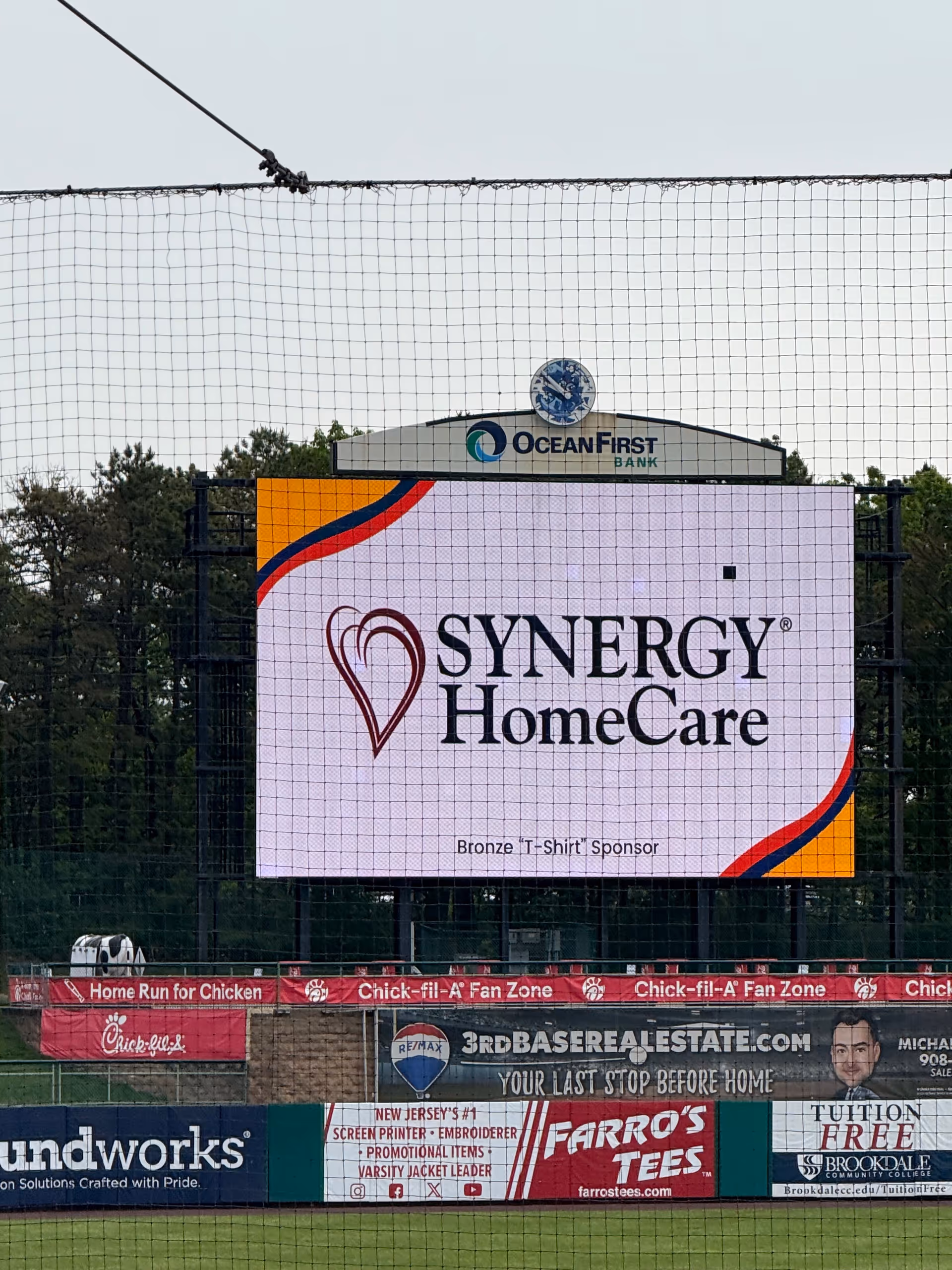 Large outdoor digital billboard displays Synergy HomeCare logo at a stadium scoreboard.
