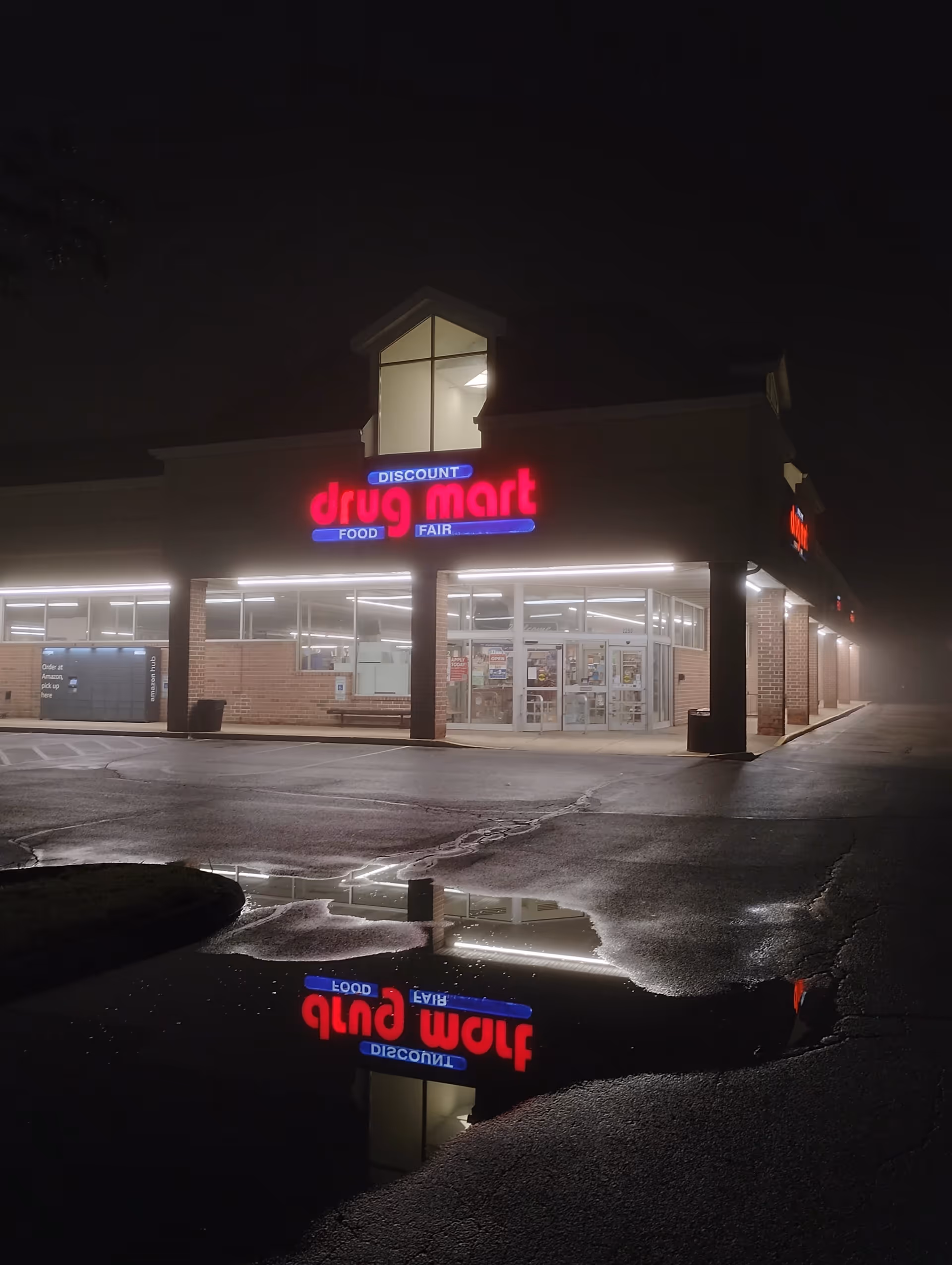 Nighttime exterior of Discount Drug Mart storefront with bright signage and puddle reflection.