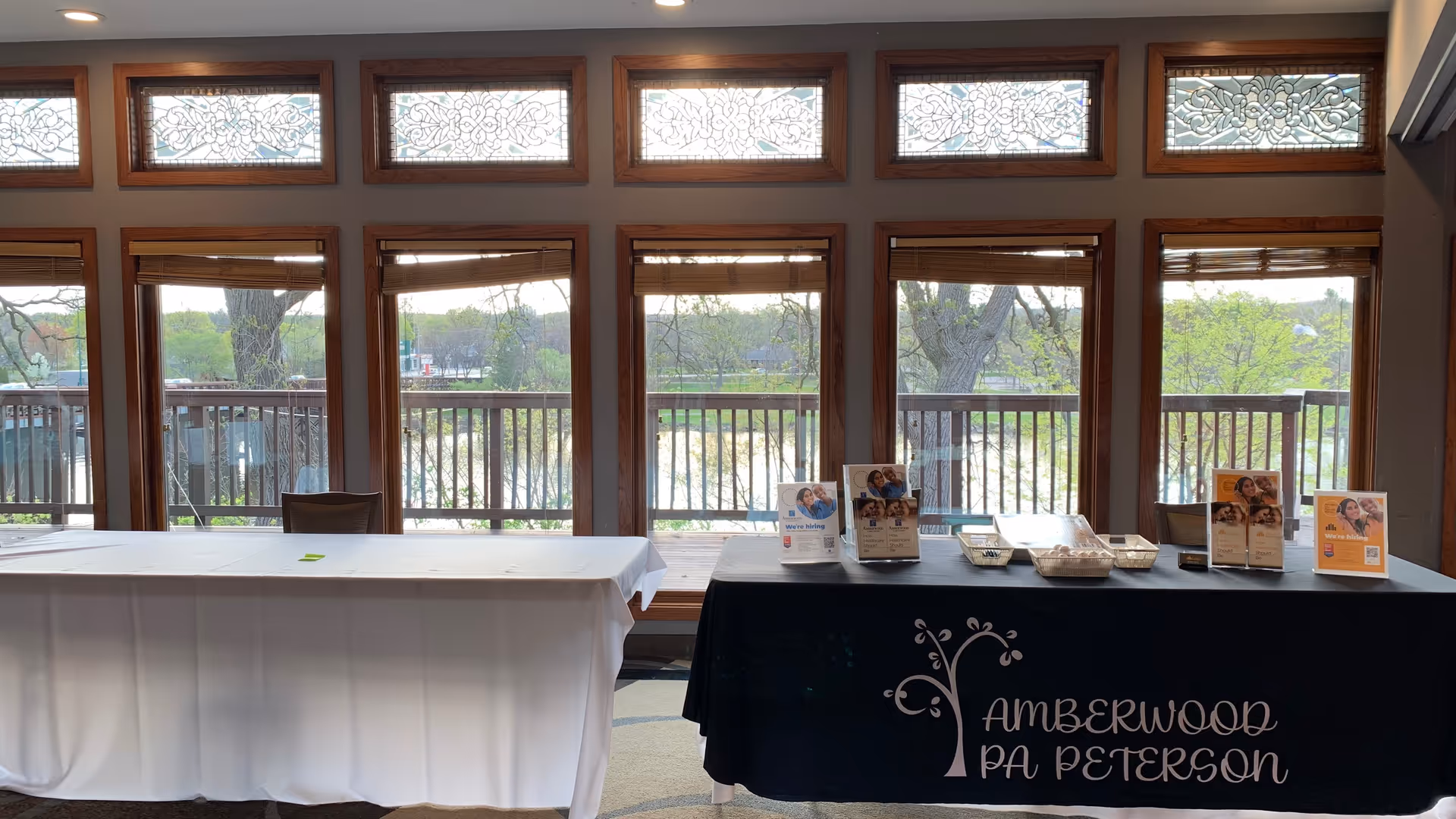 Bright indoor event setup with two long tables and brochure displays by windows.
