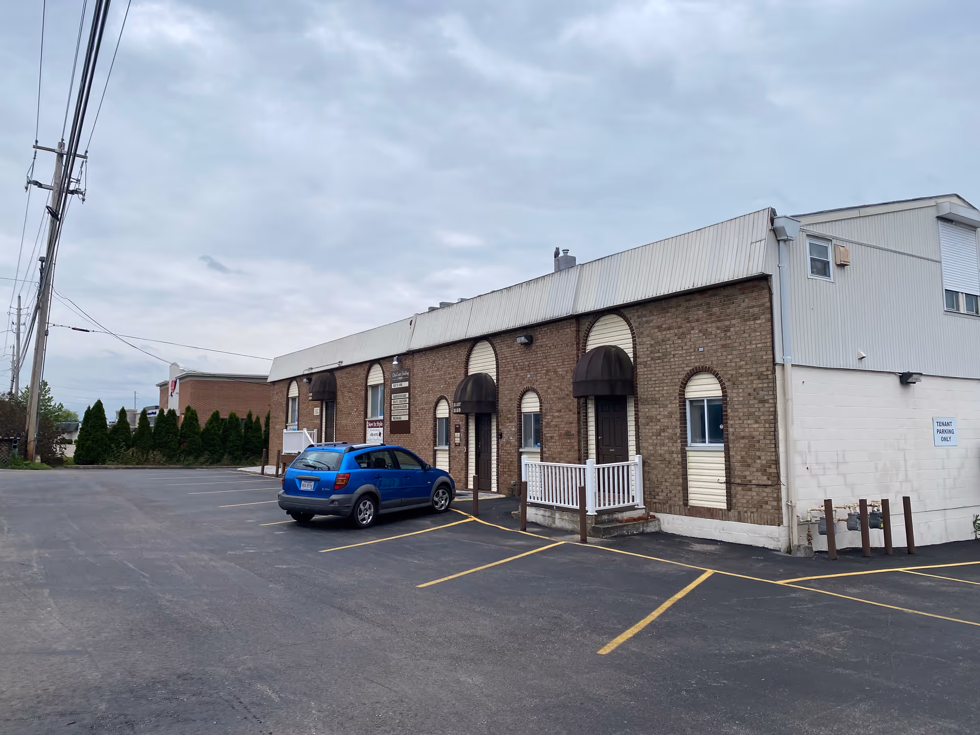 Brick facility with arched windows and awnings; blue car parked in front lot, cloudy sky.