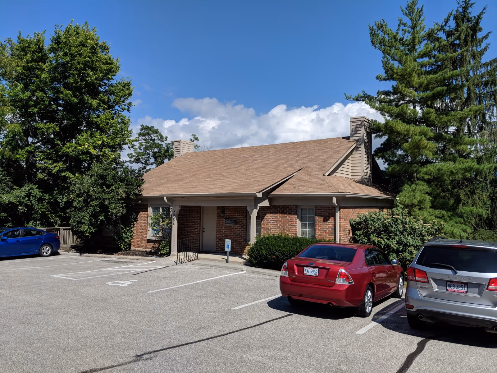 Brick senior living facility with a brown roof, parking lot, and surrounding trees.