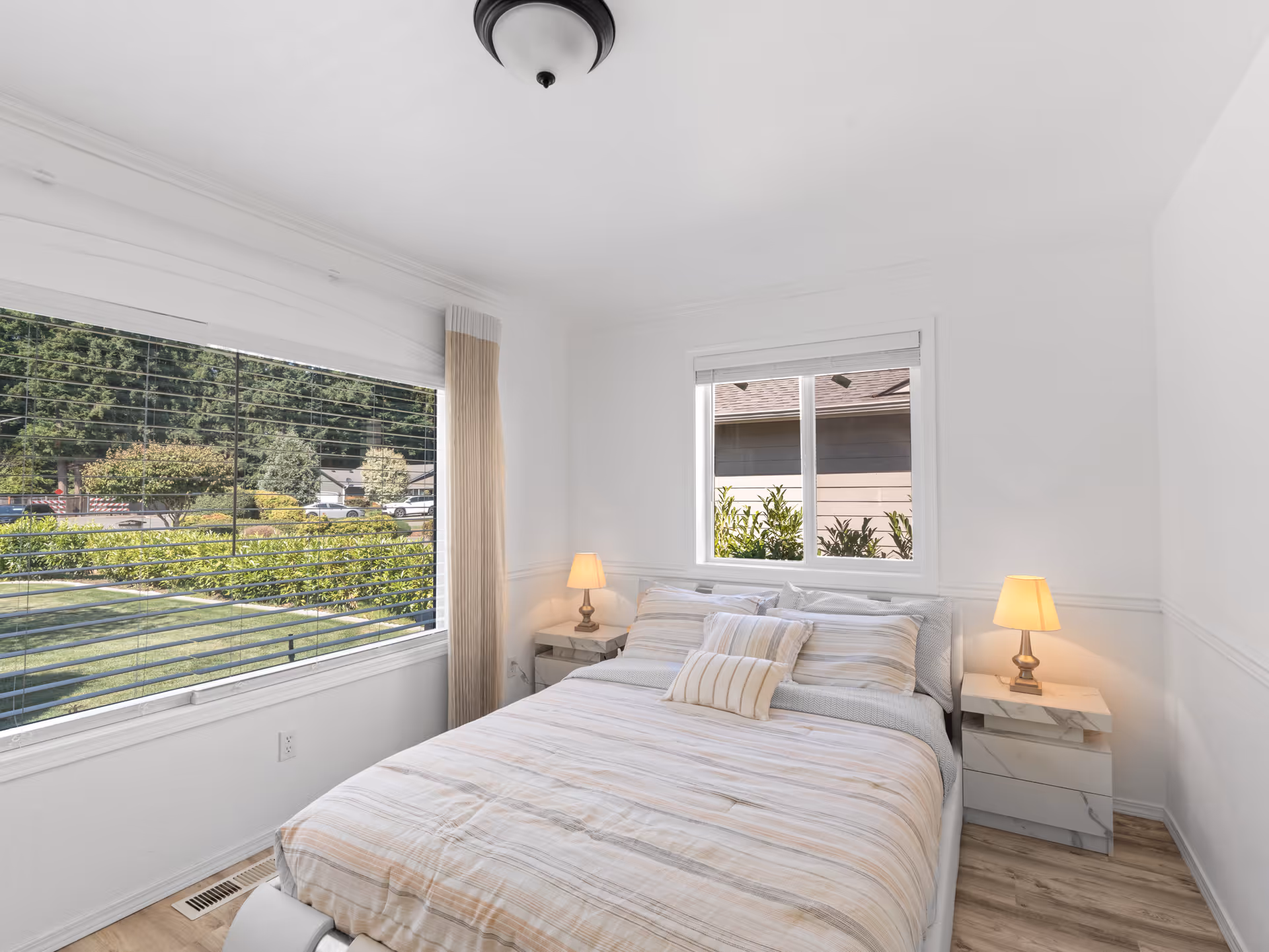 Bright bedroom with a made bed, nightstands, and a large window overlooking outdoors.