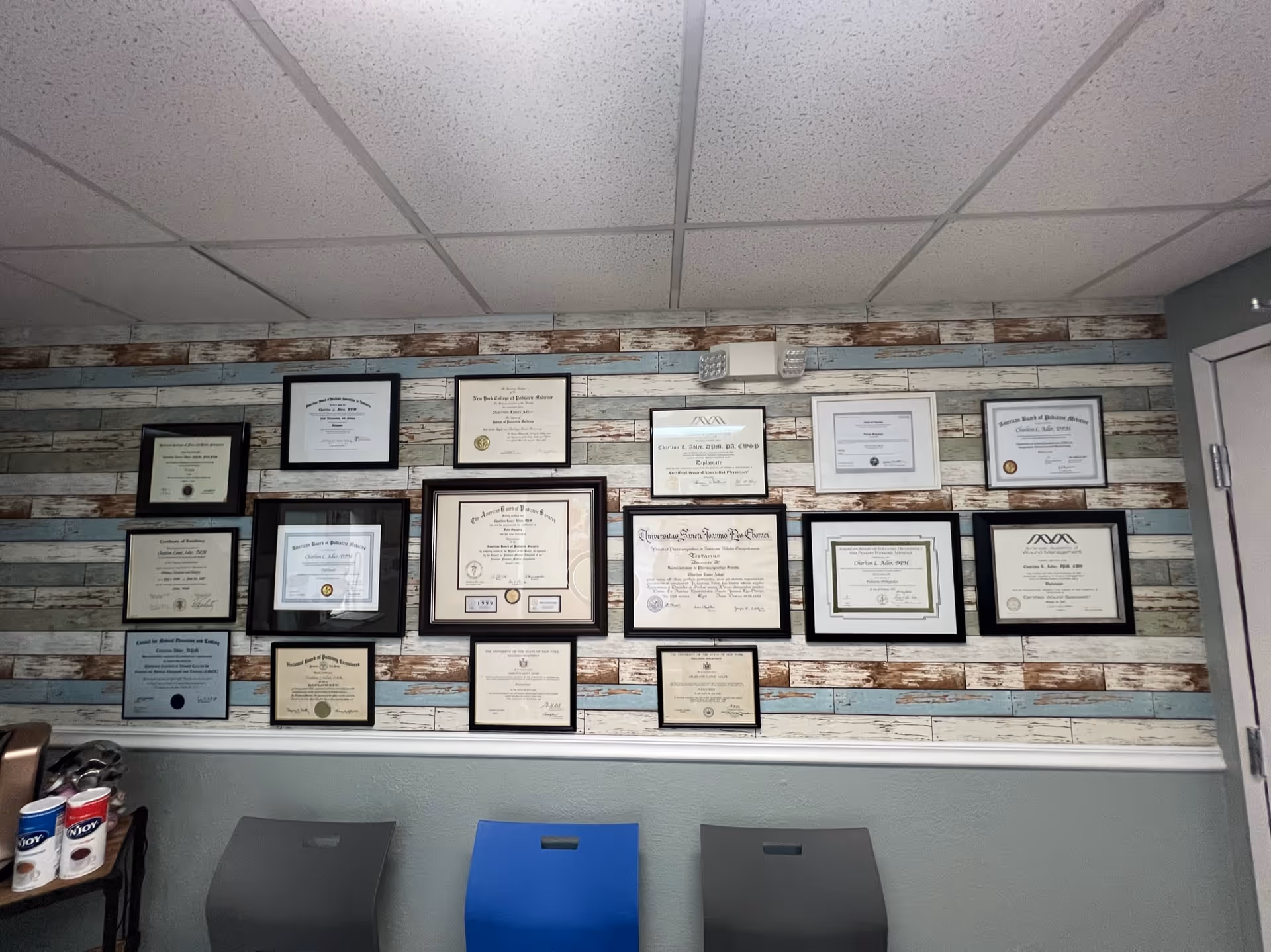 Interior wall displaying numerous framed certificates over striped wallpaper, with ceiling tiles and chairs.