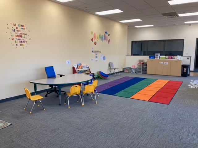 Bright interior playroom with child-sized furniture, rainbow carpet, and storage cabinets.