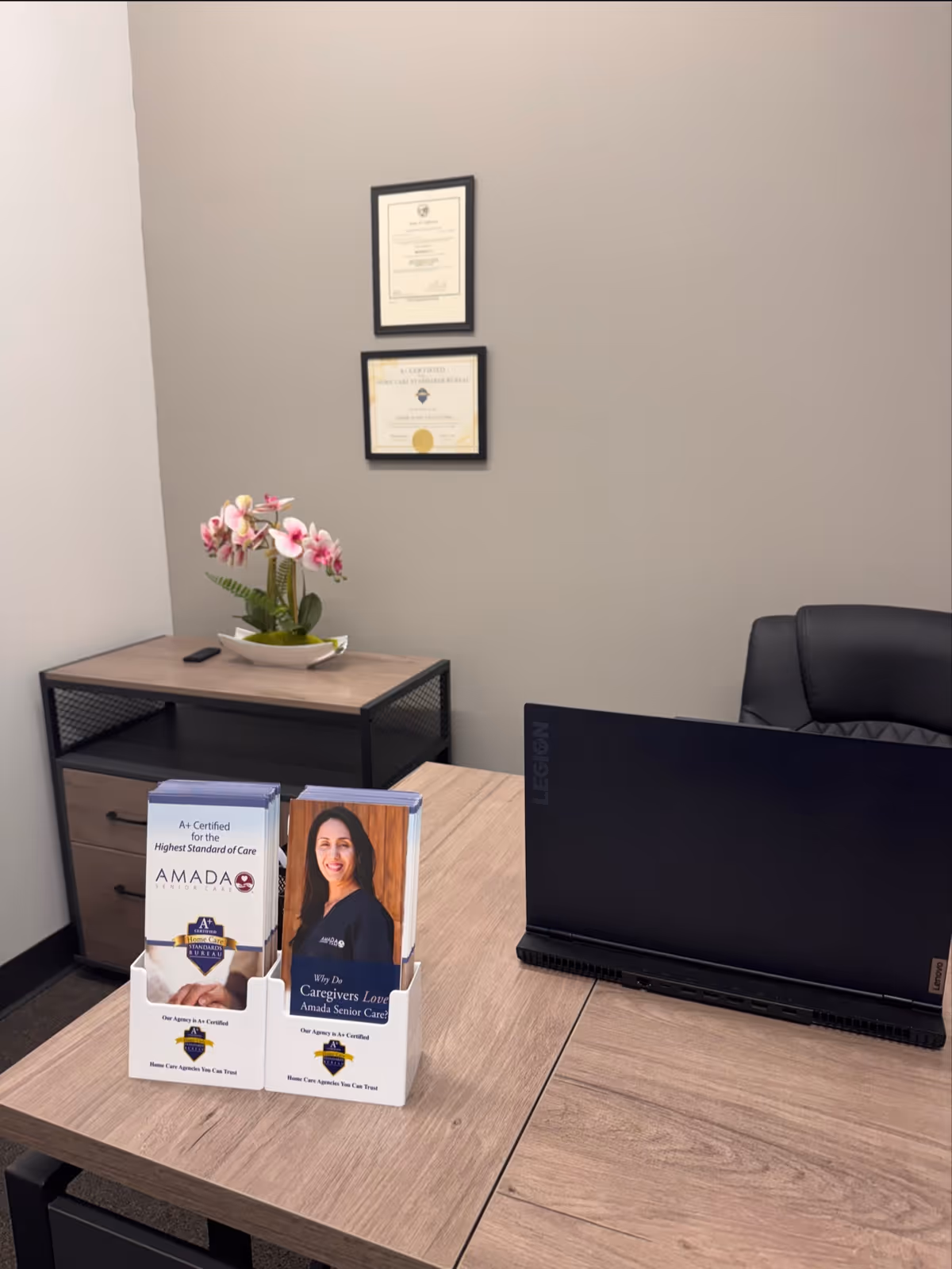 Office reception area with a desk, computer, brochure stand, orchid, and framed certificates.