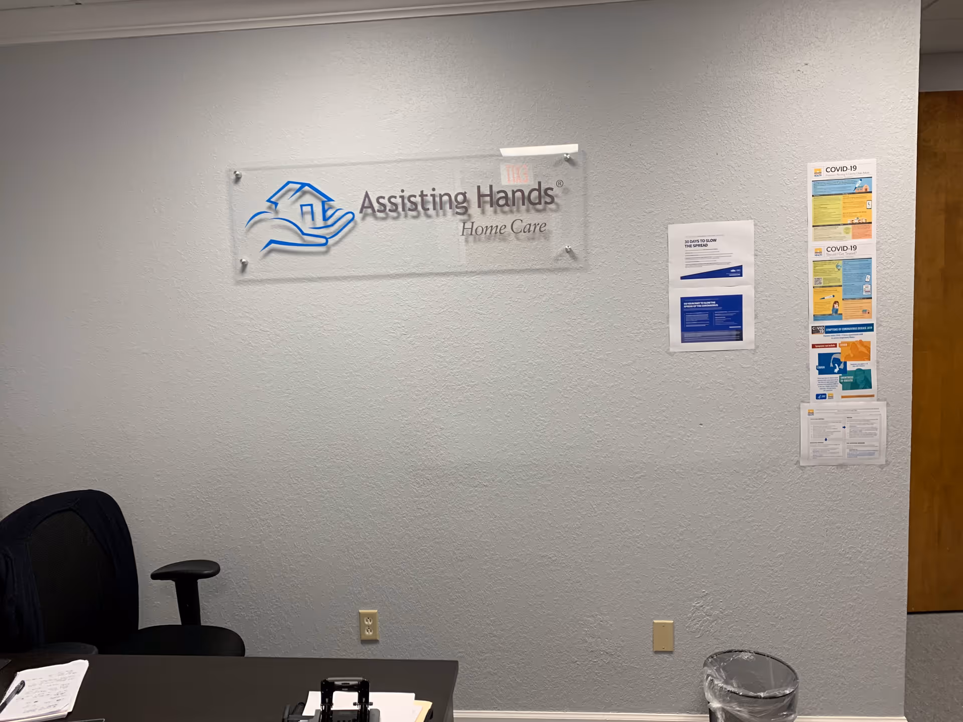 Office desk and chair against a wall with a sign for Assisting Hands Home Care.