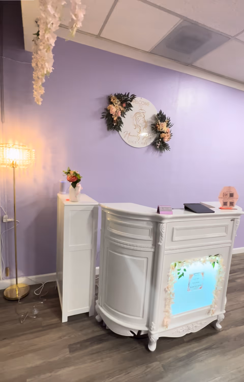 Lavender reception area with ornate white desk, floral wreaths, and warm lighting.