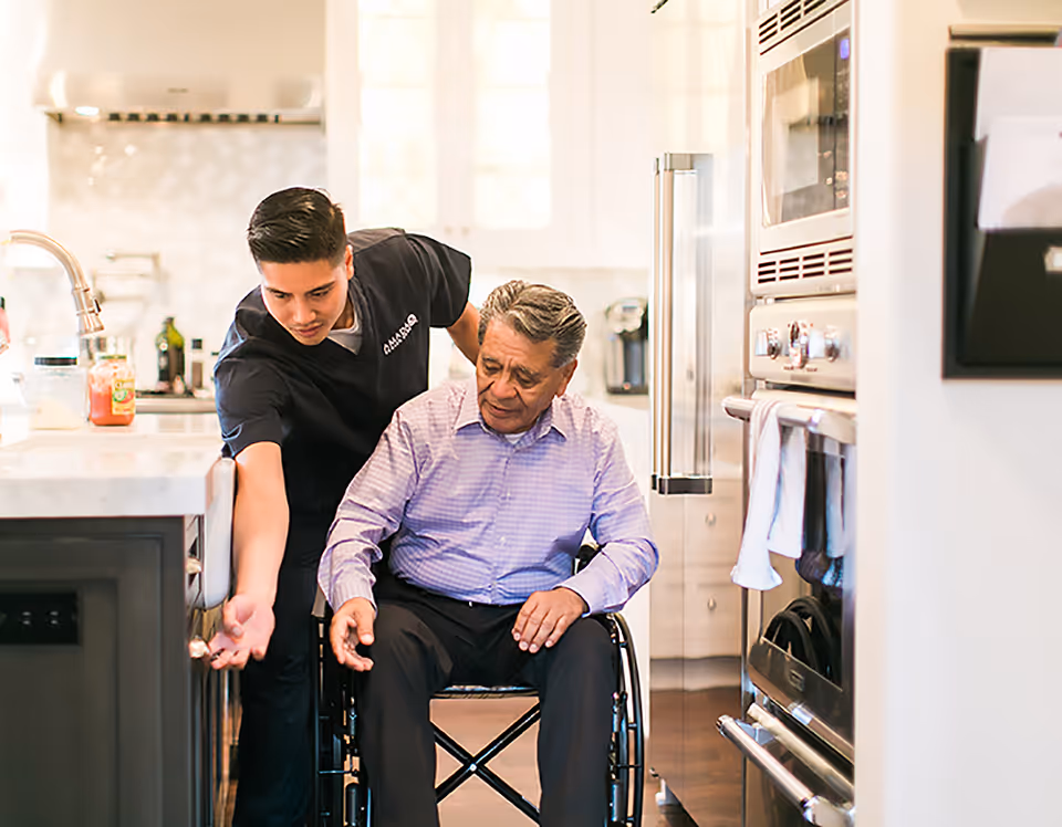 Caregiver assists an older man in a wheelchair in a bright modern kitchen.