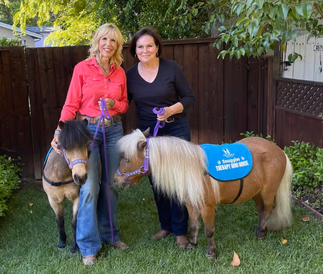 Two women stand with two miniature horses on a grassy backyard.