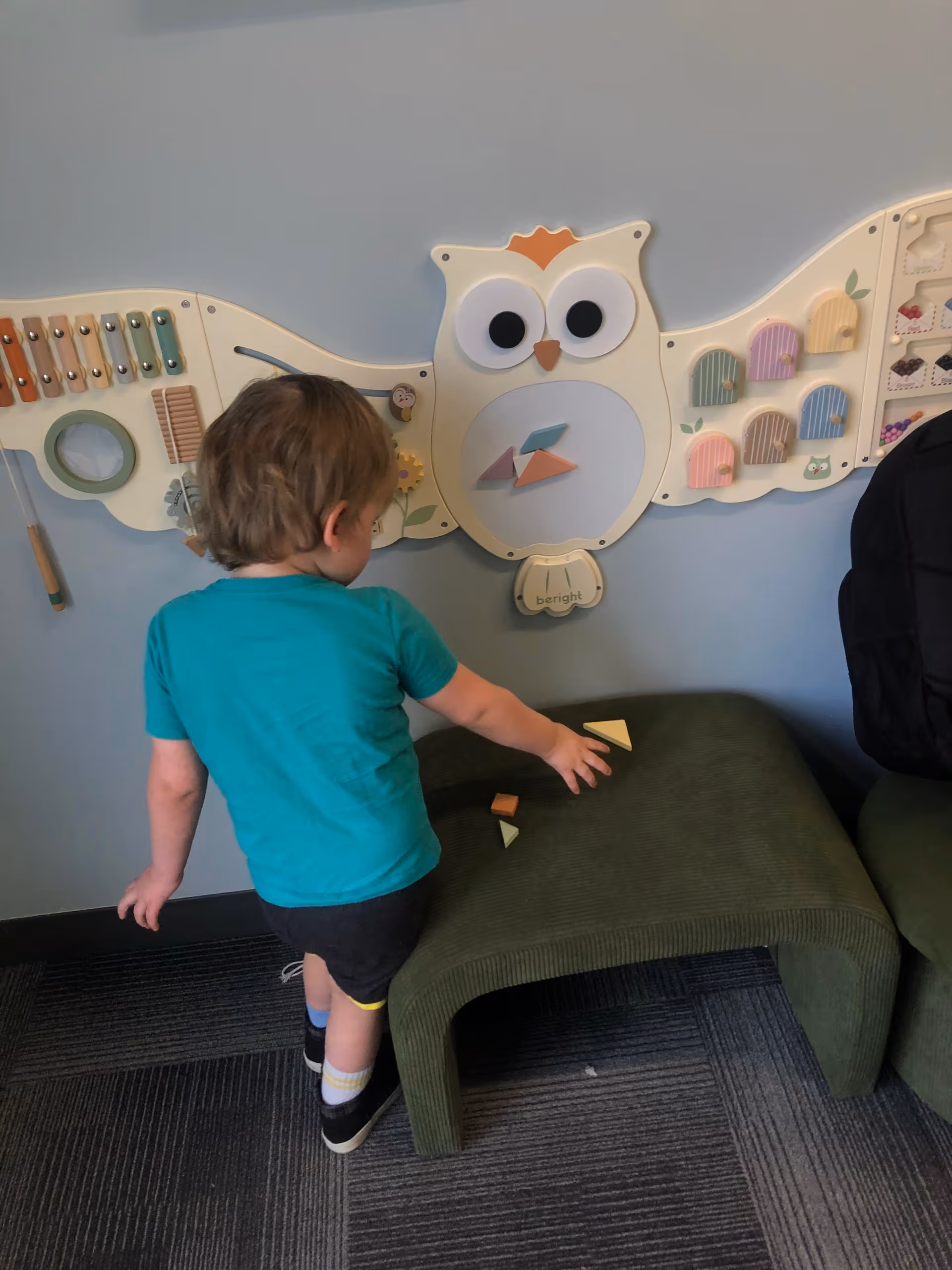 A child in teal shirt plays with an owl-themed wall activity panel indoors.