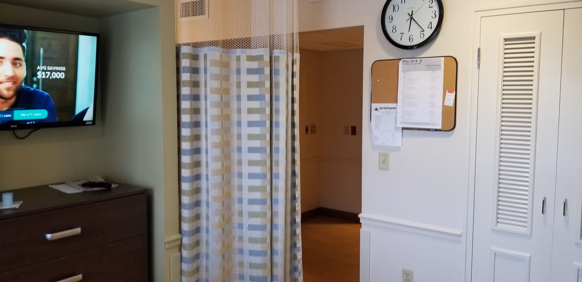 TV mounted on wall beside dresser, striped curtain partitions hallway in a Miami Jewish Health facility.
