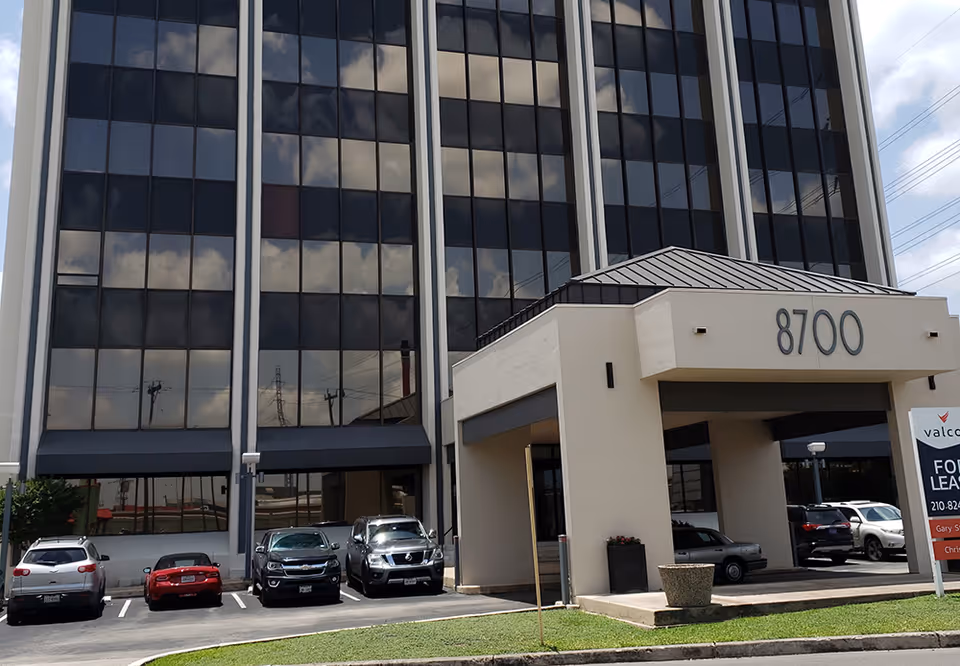 Tall glass-front building with parked cars outside the entrance marked 8700