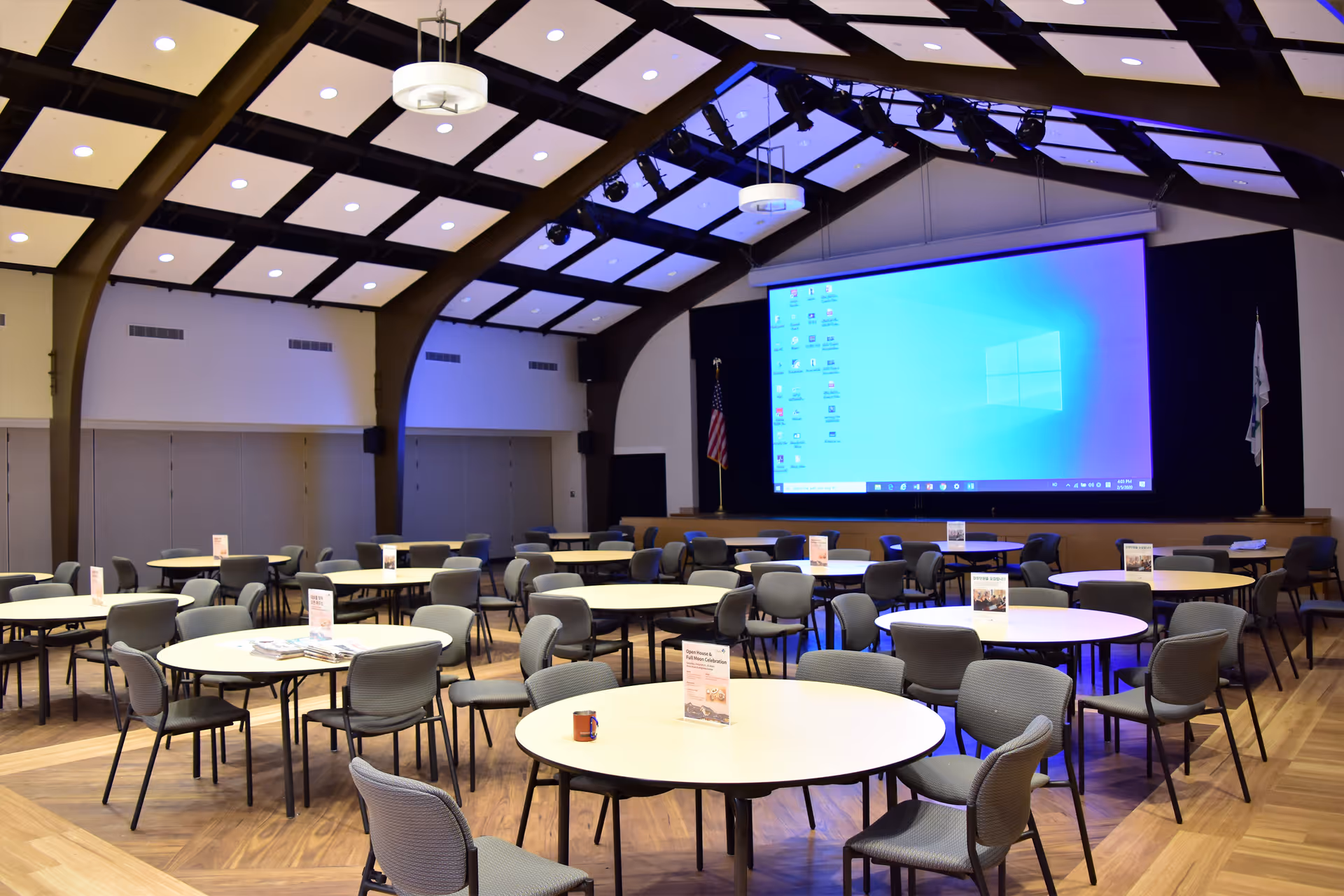 Large interior hall with round tables, chairs, and a large projector screen.