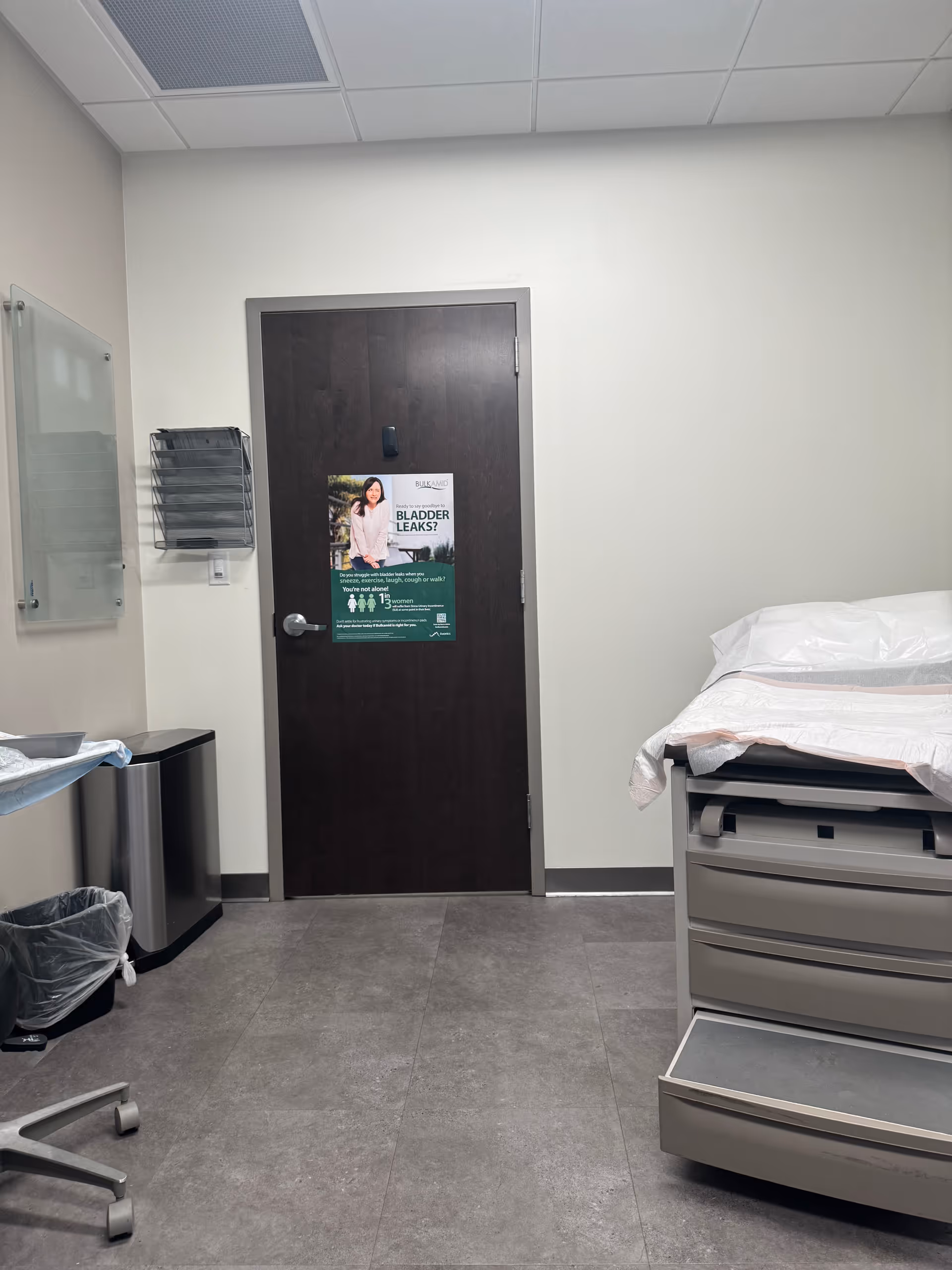 Clinical examination room with a bed, a closed door, and medical equipment along the wall.