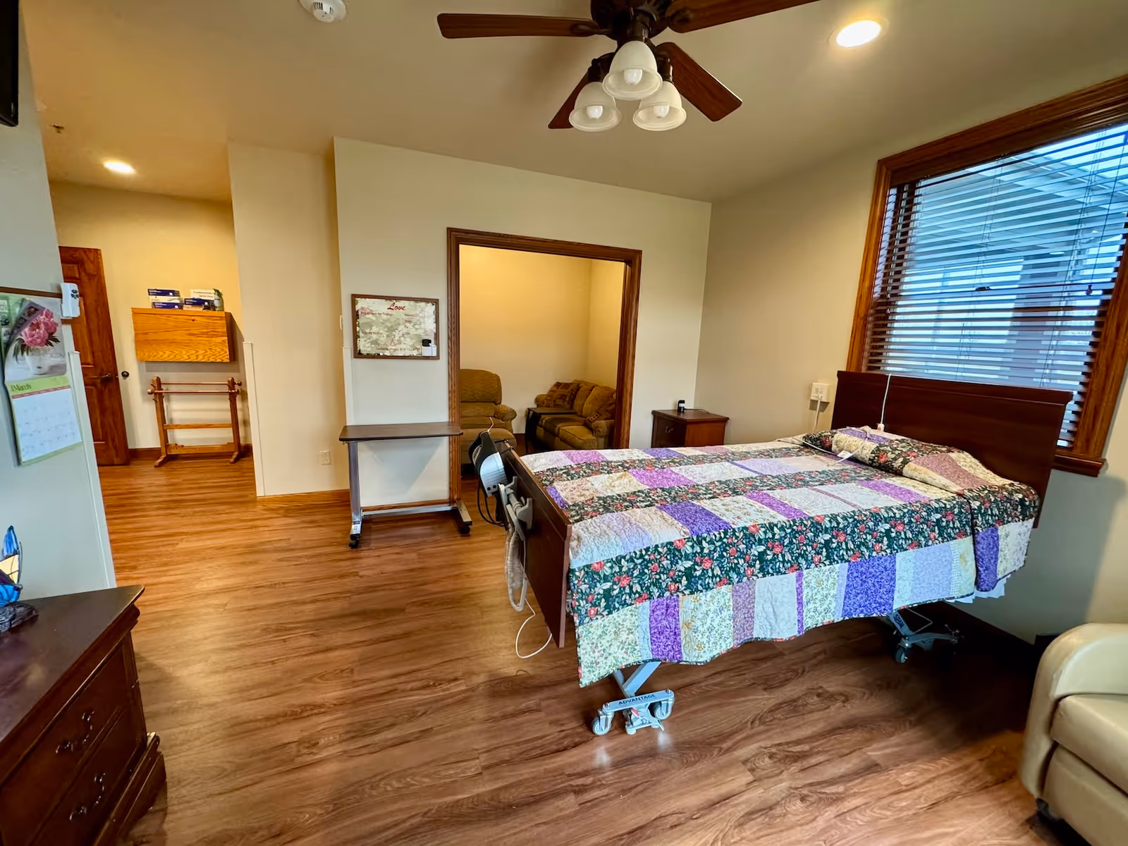 A furnished bedroom with patchwork quilted bed, wooden floors, ceiling fan, and blinds.