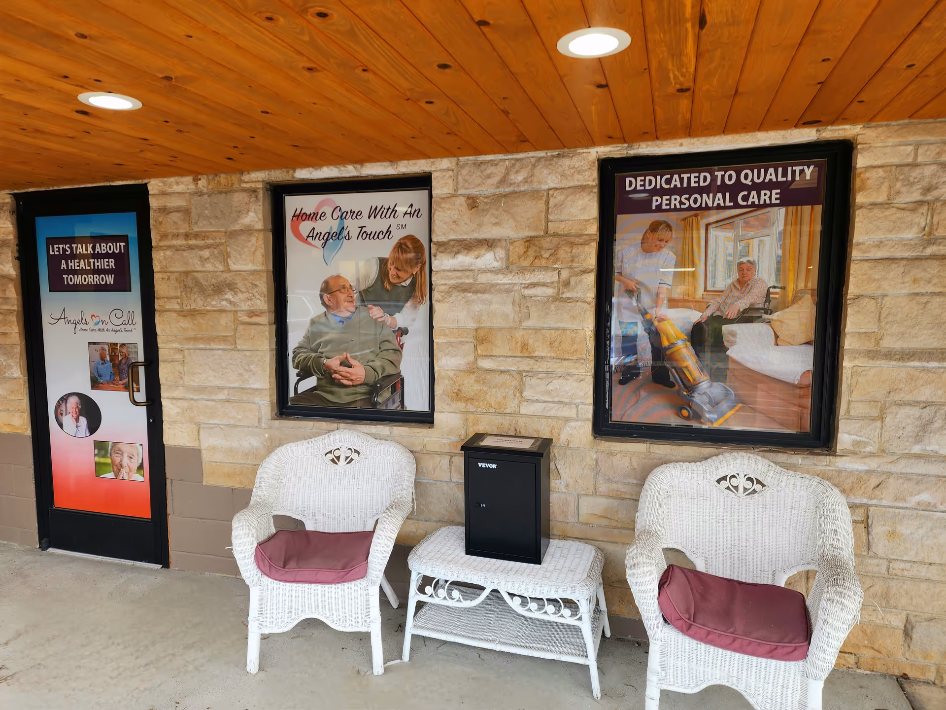 Exterior entry with stone wall, wooden canopy, two white wicker chairs, and posters.