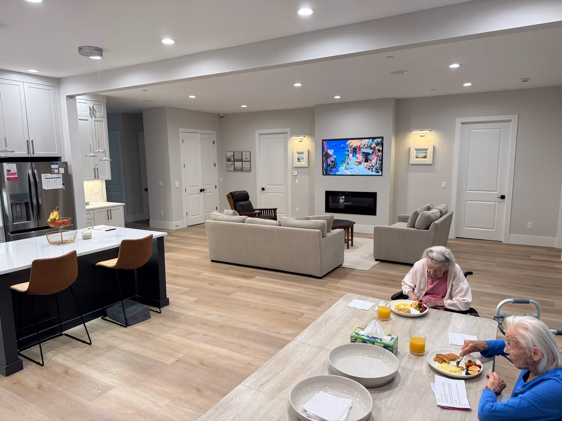 Bright open common living area with sofas, kitchen island, and two seniors dining.