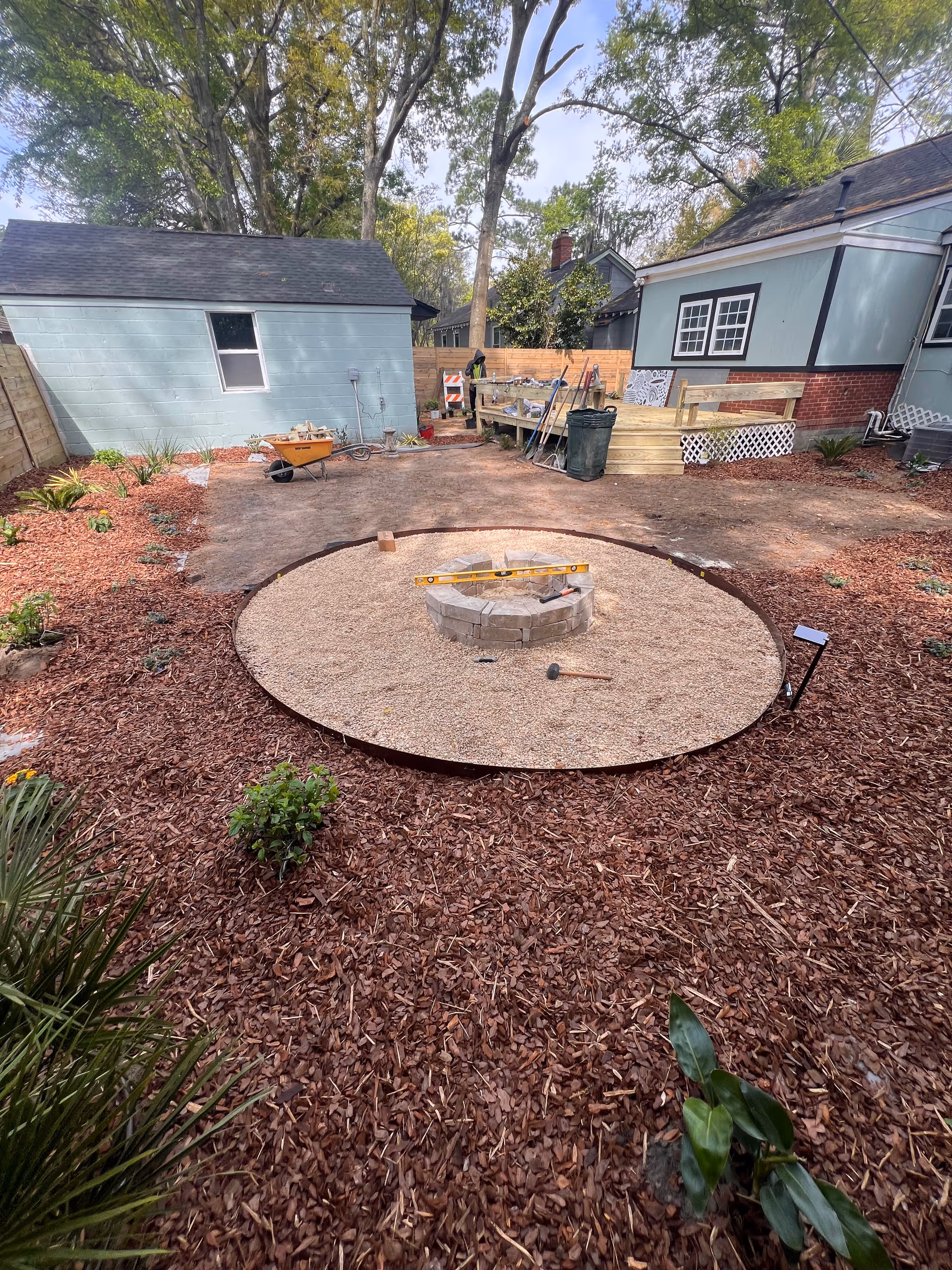 Backyard under construction: circular fire pit area with gravel, mulch, tools, and a decked house.