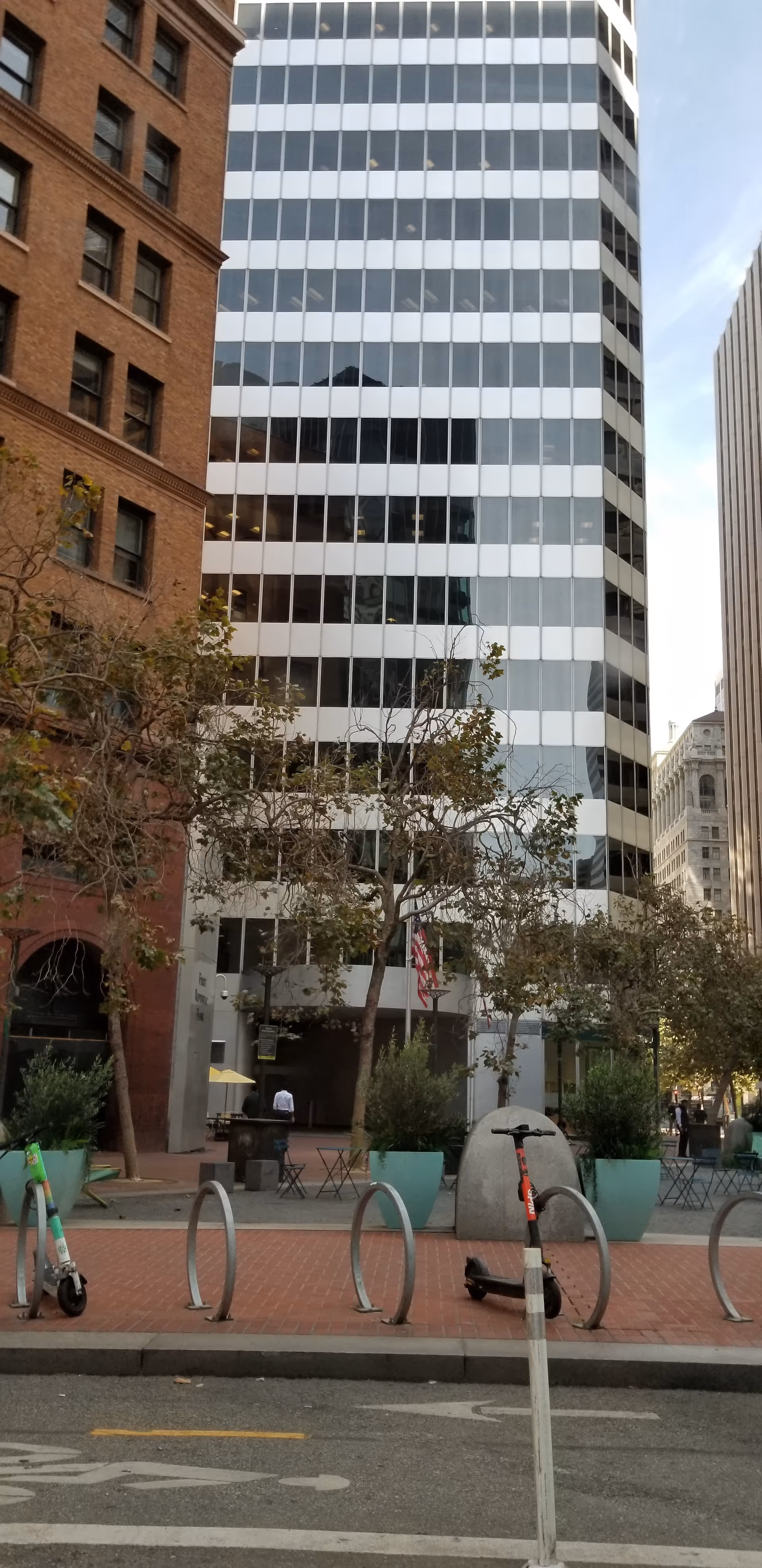 Urban street scene with towering glass buildings, planters, trees, and outdoor seating.